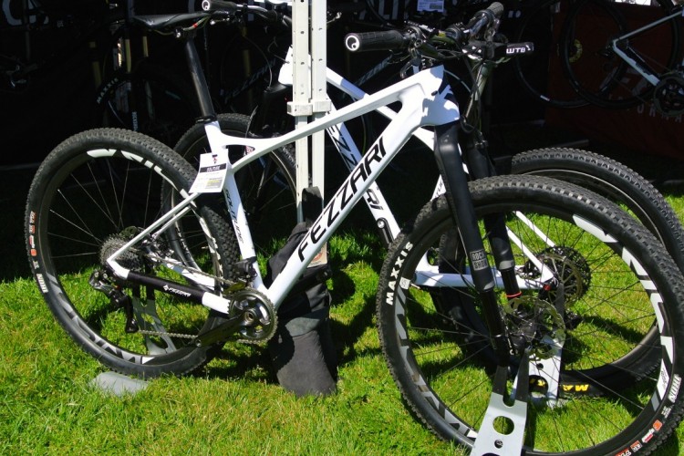 A white mountain bike with a sleek design, featuring wide tires and a front suspension fork. The bike is positioned on grass with other bikes visible in the background, indicating a display or event setting.