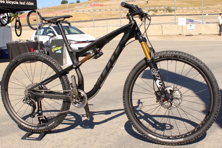 A high-performance mountain bike with a sleek black frame and gold suspension components, positioned on a concrete surface. The bike features wide tires with knobby tread for traction, a drivetrain with visible gears and chain, and disc brakes. In the background, a tent displays signage for free bicycle test rides, with additional bicycles and a parked car nearby. The setting suggests an outdoor event or cycling exhibition.