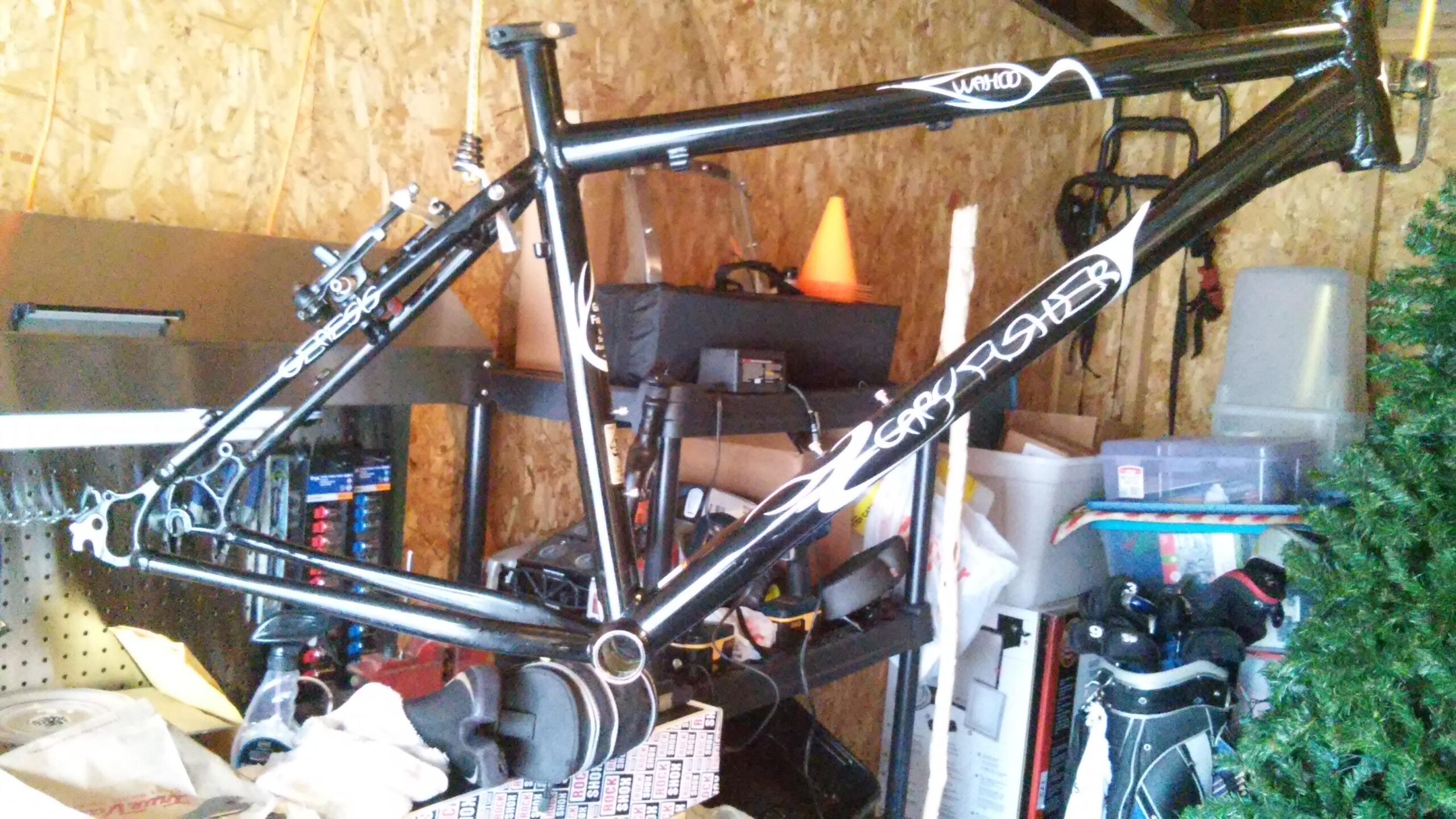 Gary Fisher Wahoo: A black bicycle frame is suspended in a cluttered garage, surrounded by various items including boxes, golf clubs, and a Christmas tree. The frame features white detailing and branding. The background showcases tools and storage containers, illustrating a typical home workspace.