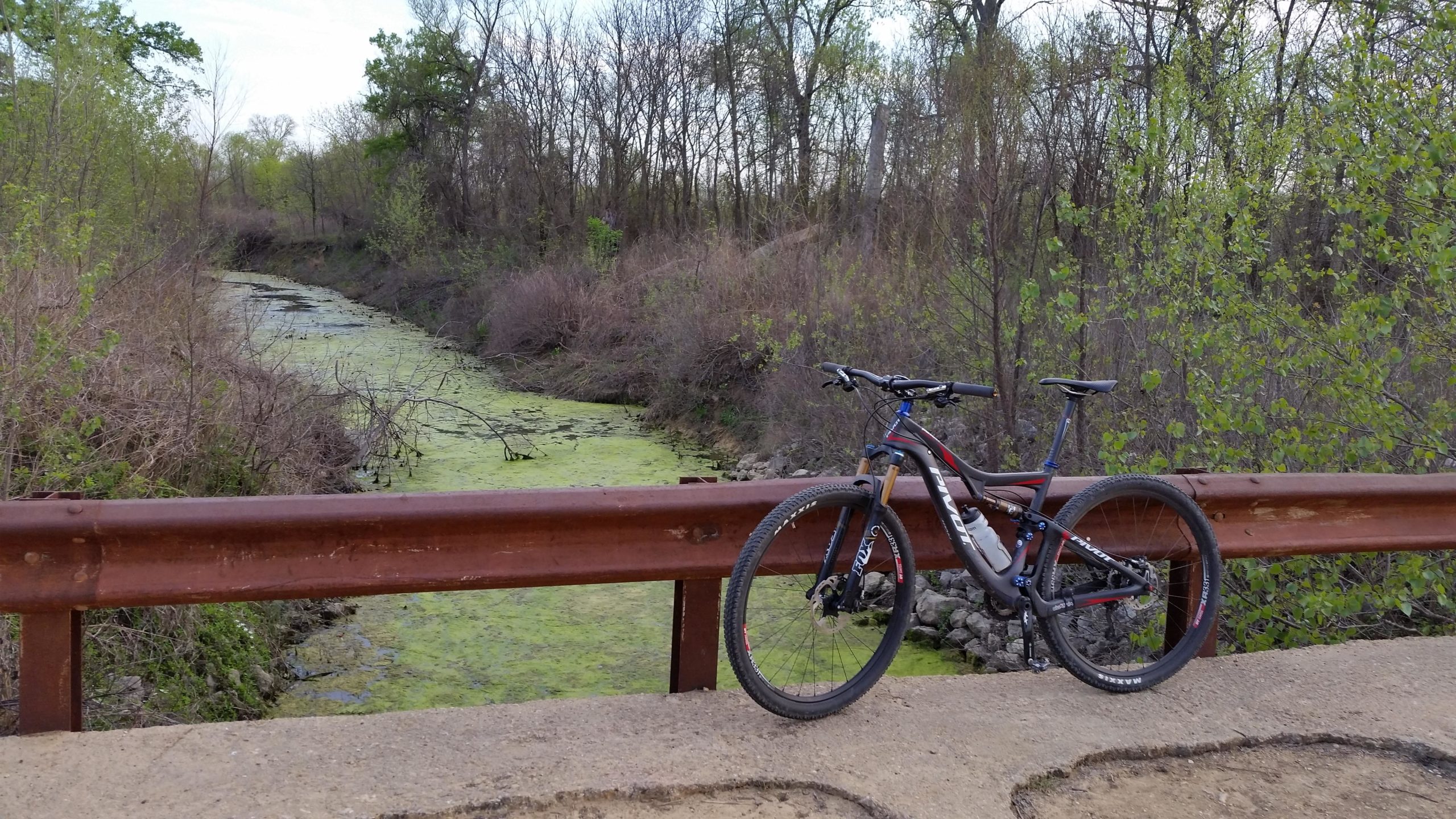 A mountain bike leans against a rusted railing, overlooking a narrow, algae-covered stream surrounded by sparse vegetation and trees in early spring. Knob Hills Grapevine Lake mountain bike trail.