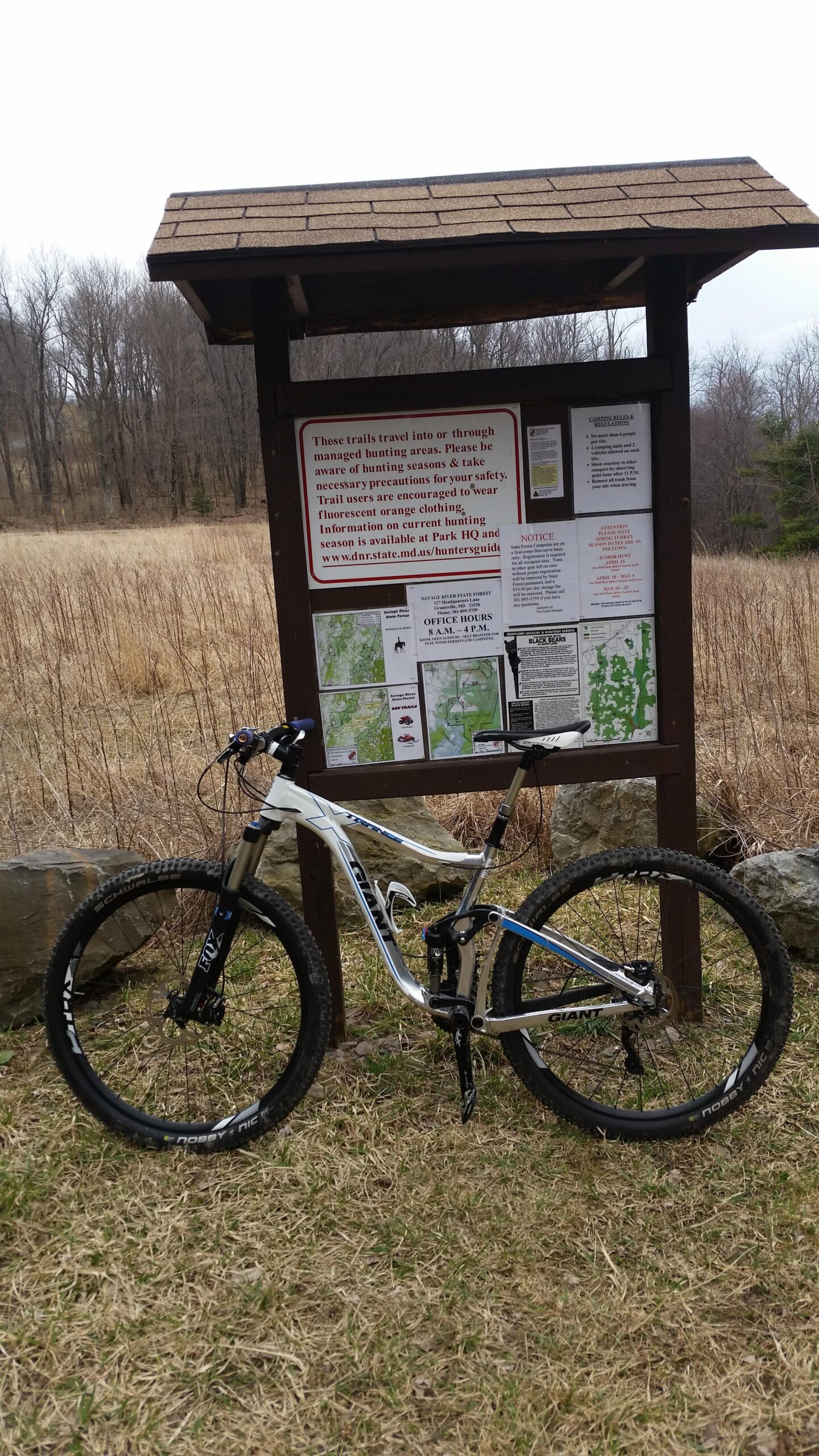 Motobecane NightTrain Bullet: A mountain bike is parked beside an information sign at a trailhead, surrounded by grassy fields and bare trees. The sign includes safety information about hunting seasons and trail maps.