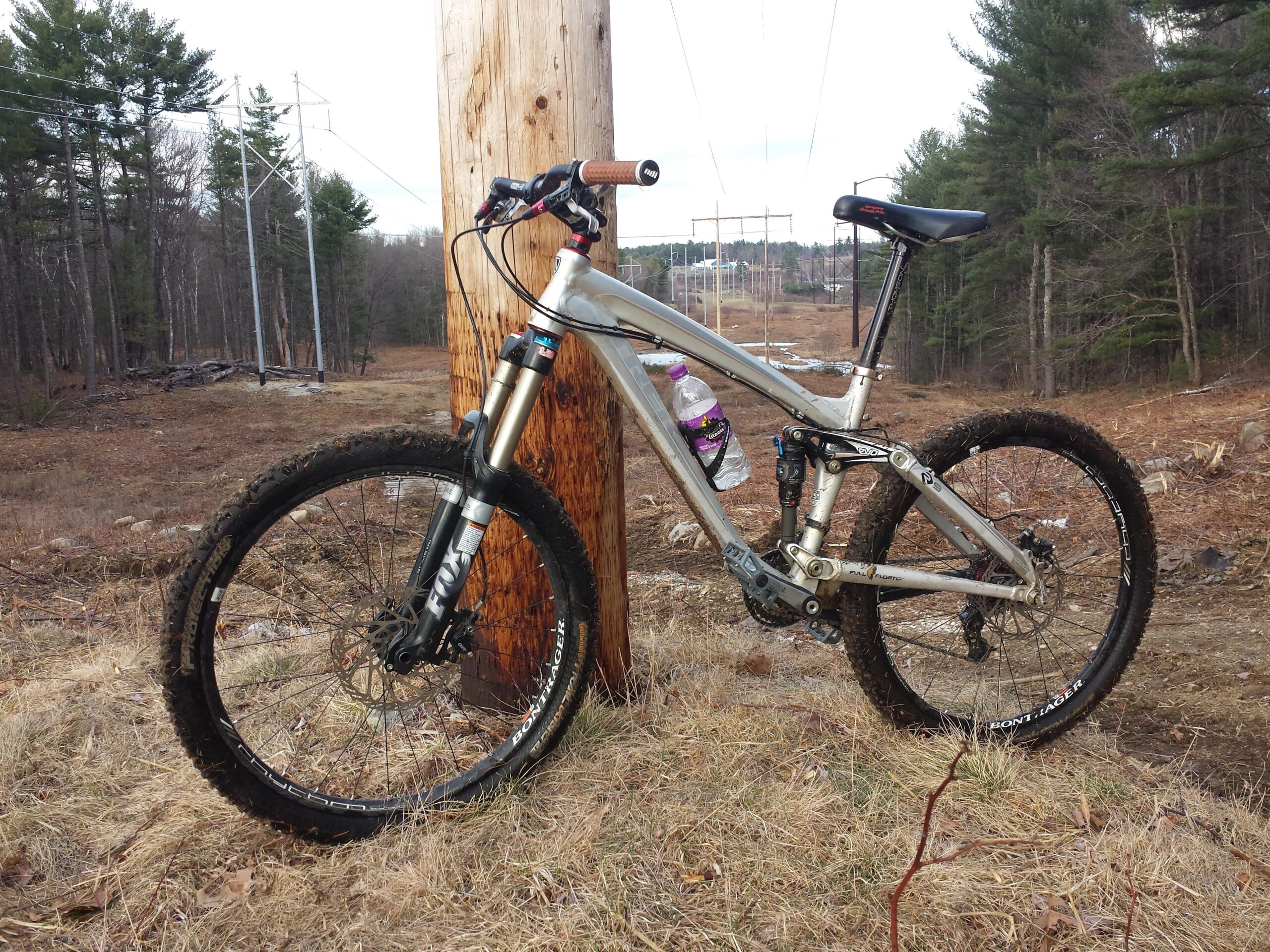 Trek Remedy 9: A silver mountain bike leaning against a wooden utility pole, with a water bottle attached to the frame. The bike is positioned on a grassy area with some dirt and gravel, surrounded by a sparse forest of pine trees. In the background, power lines stretch across an open field, indicating a rural outdoor setting.