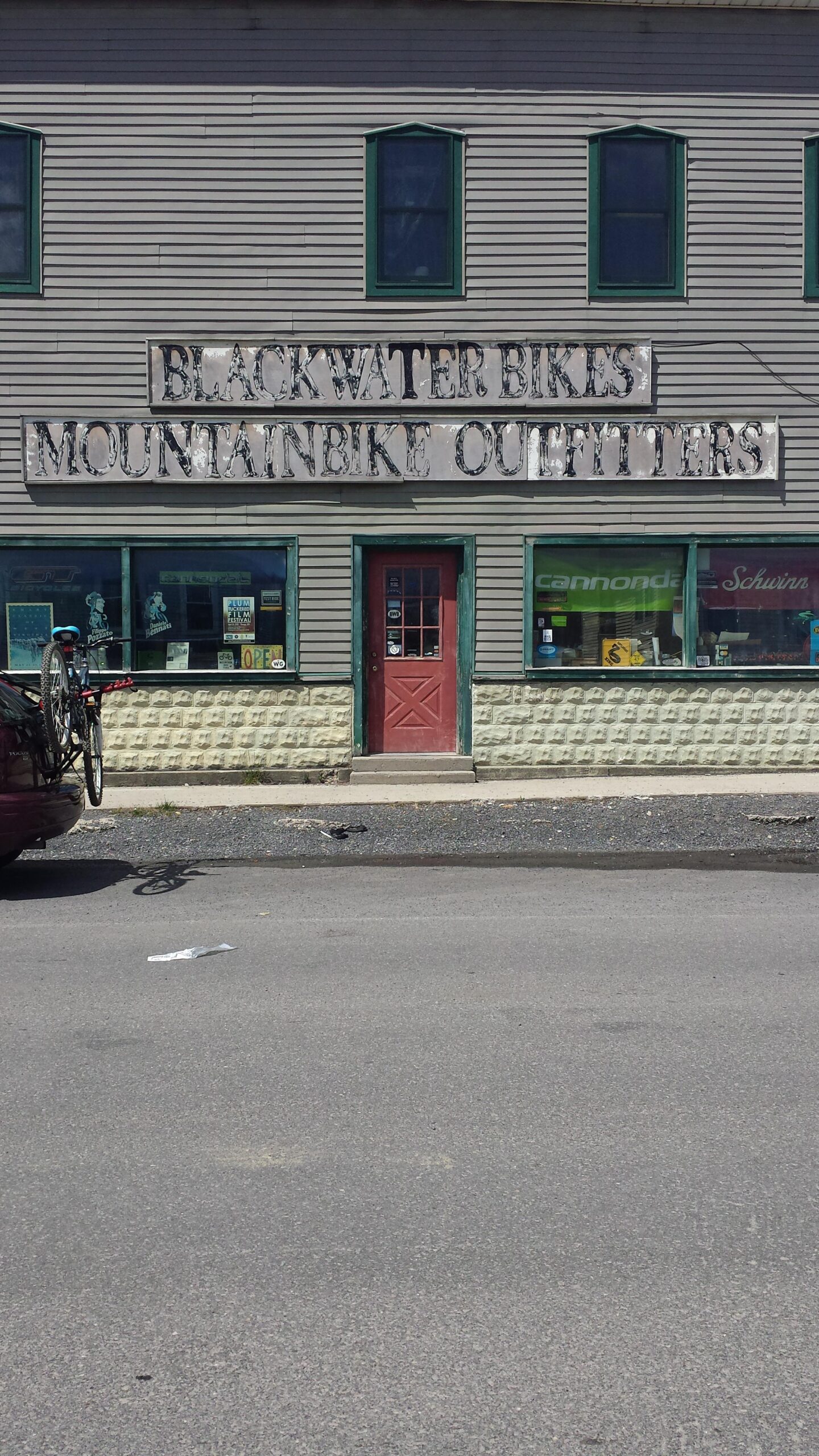 Image of a bike shop storefront featuring a large sign that reads "Blackwater Bikes Mountainbike Outfitters." The building has green-trimmed windows and a red door, with a bicycle mounted on a car parked in front. Various signs and advertisements can be seen in the windows.