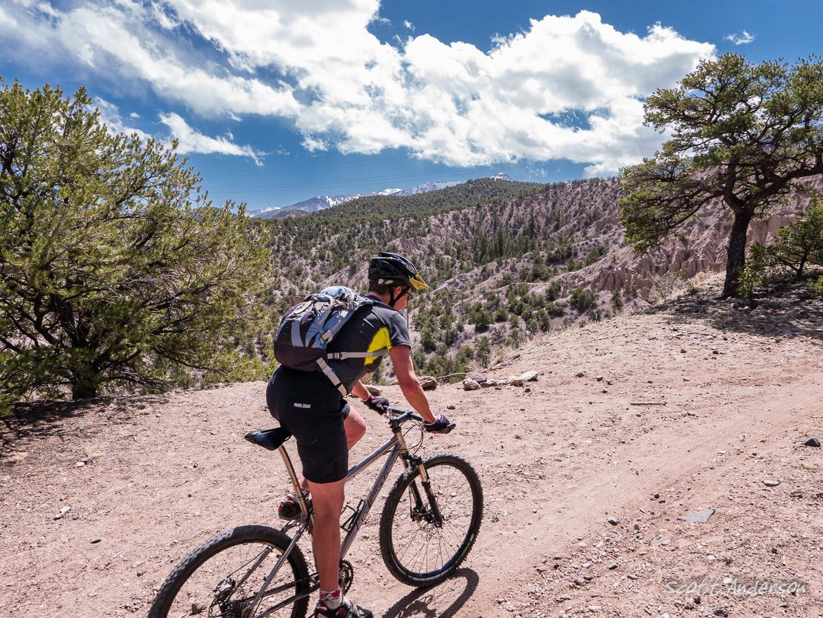 A mountain biker navigates a dirt trail surrounded by scenic hills and trees under a partly cloudy sky. The cyclist is wearing a helmet and a backpack, focusing on the path ahead as they ride through a natural landscape. Race Track mountain bike trail.