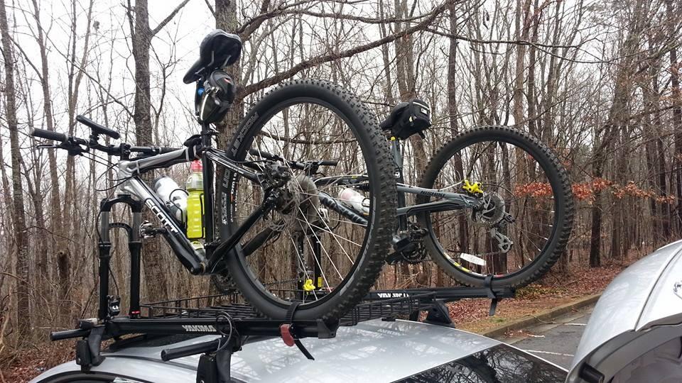 Scott Reflex 20: Two mountain bikes secured on the roof rack of a car, with trees in the background. One bike has a water bottle attached to it. The scene is set in a wooded area, indicating an outdoor activity.