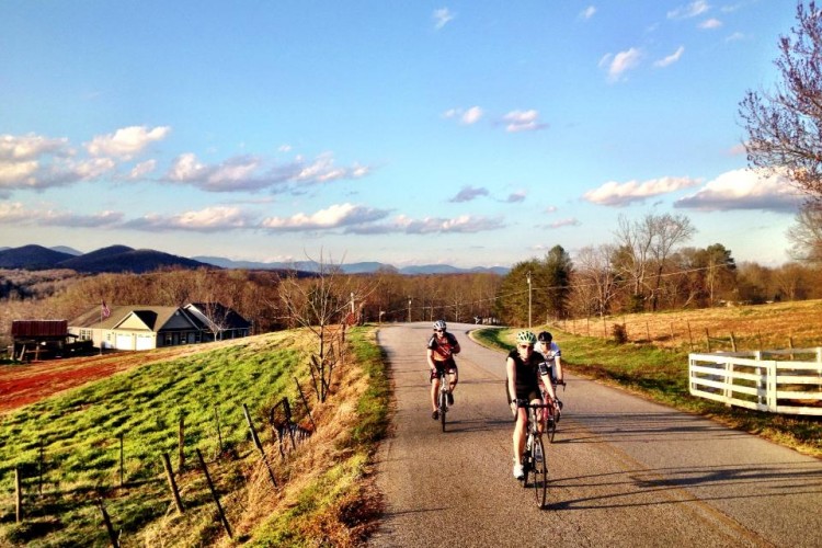 Three cyclists riding along a winding road through a scenic landscape with rolling hills, green fields, and a farmhouse. The sky is clear with fluffy clouds, and mountains can be seen in the distance, indicating a sunny day.