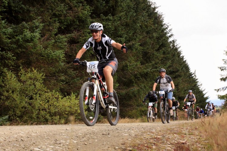 A group of mountain bikers ride along a gravel path surrounded by trees. One cyclist in the foreground is smiling and giving a thumbs up while wearing a helmet and cycling gear, with a race number on their bike. Other cyclists can be seen in the background, creating a dynamic scene of outdoor activity.