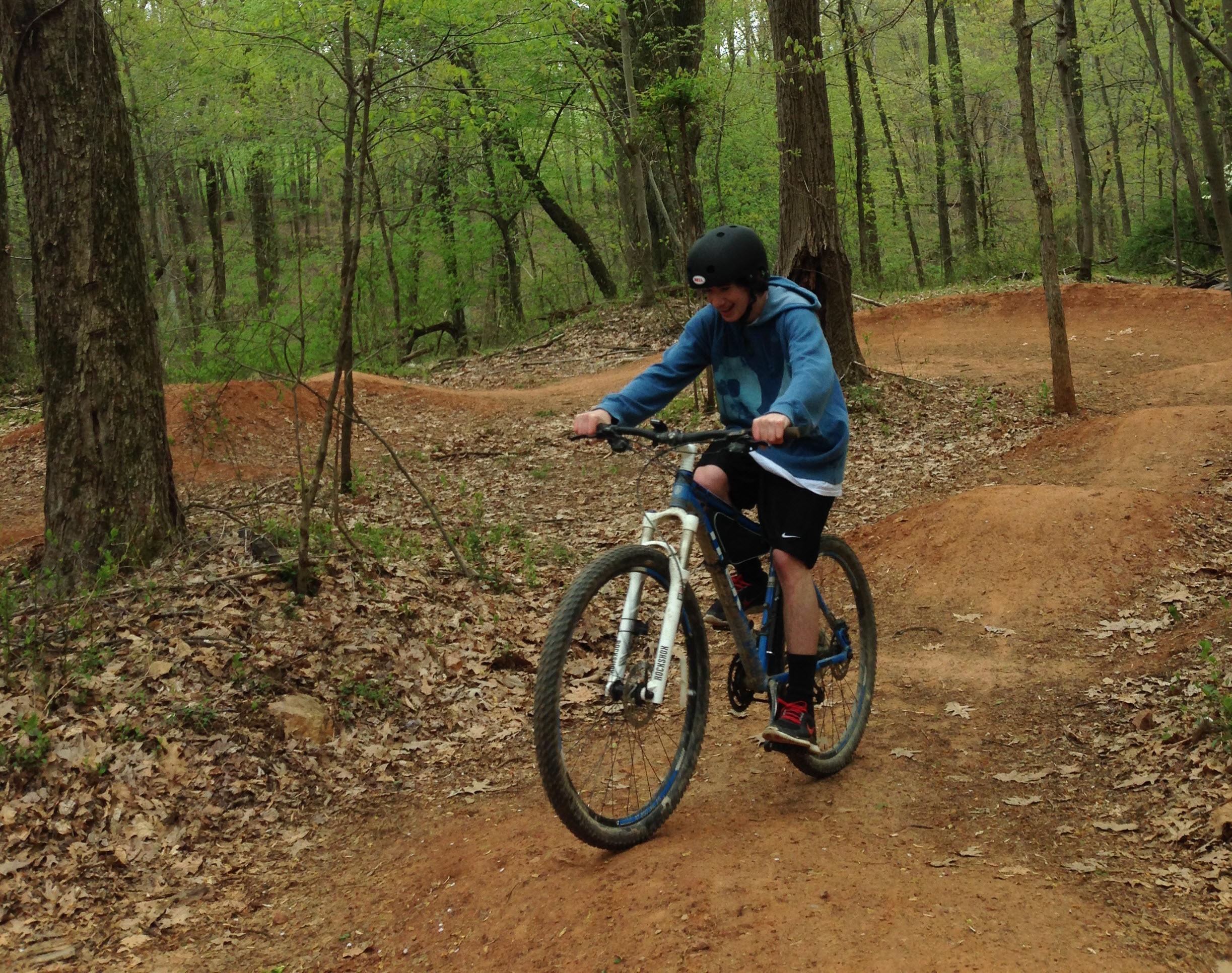 A young person wearing a helmet is riding a mountain bike on a dirt trail surrounded by trees. The trail has small jumps and is covered with fallen leaves, indicating an outdoor biking park. The scene conveys a sense of adventure and fun in a natural setting. Freedom Center mountain bike trail.