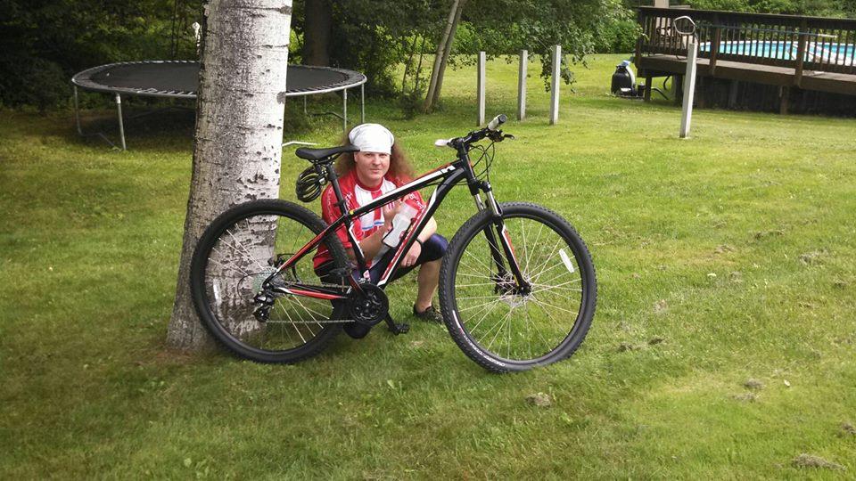 Specialized Hardrock Sport Disc 29er: A person with long hair sits beside a mountain bike leaning against a tree in a grassy yard. In the background, there is a trampoline and a deck with a swimming pool, surrounded by trees. The setting appears to be a sunny, relaxed outdoor environment.