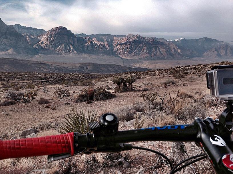 A mountain biking view overlooking a dusty landscape with rugged mountains in the background. The foreground features a close-up of the bike handlebar with red grips, while desert vegetation is scattered in the arid terrain. Cloudy skies loom above, hinting at a dramatic backdrop. Cowboy Trails mountain bike trail.