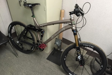 Ellsworth Moment sst.2: Mountain bike leaning against a wall in an indoor space, featuring a lightweight frame, high-performance tires, and suspension forks. The bike has a sleek design with black and silver elements, red pedals, and is positioned near a filing cabinet.
