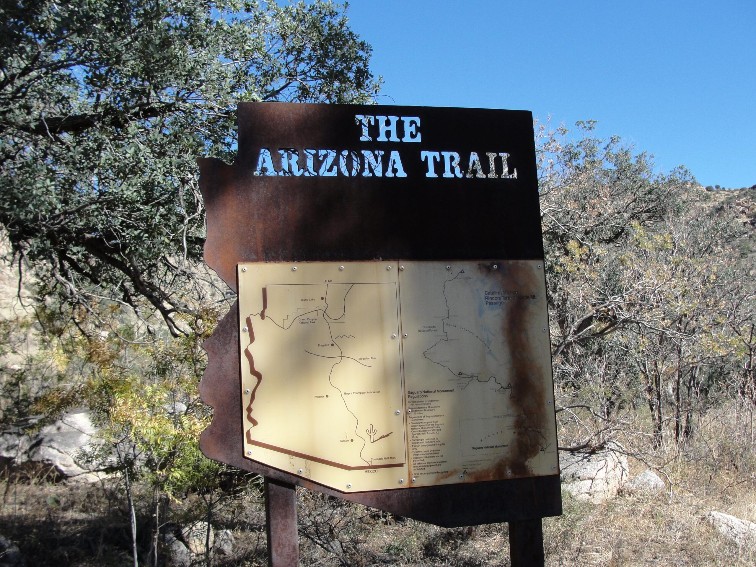 Sign for the Arizona Trail, featuring a map of the trail route. The sign is made of metal, with the title "THE ARIZONA TRAIL" displayed at the top. Surrounding foliage and a clear blue sky are visible in the background. Arizona Trail mountain bike trail.