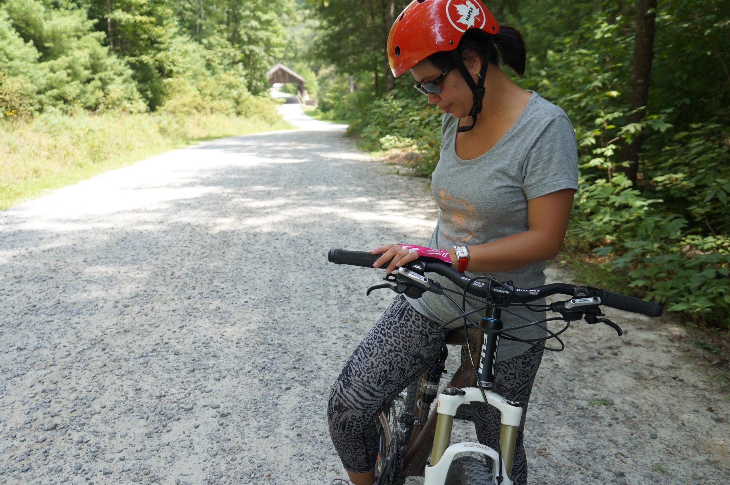 A person wearing a bright orange helmet, sunglasses, and athletic clothing is standing next to a mountain bike on a gravel path surrounded by greenery. They appear to be checking their bike or a device on their wrist, focusing intently while the sunlight filters through the trees in the background. A rustic shelter can be seen in the distance along the path. DuPont State Forest mountain bike trail.