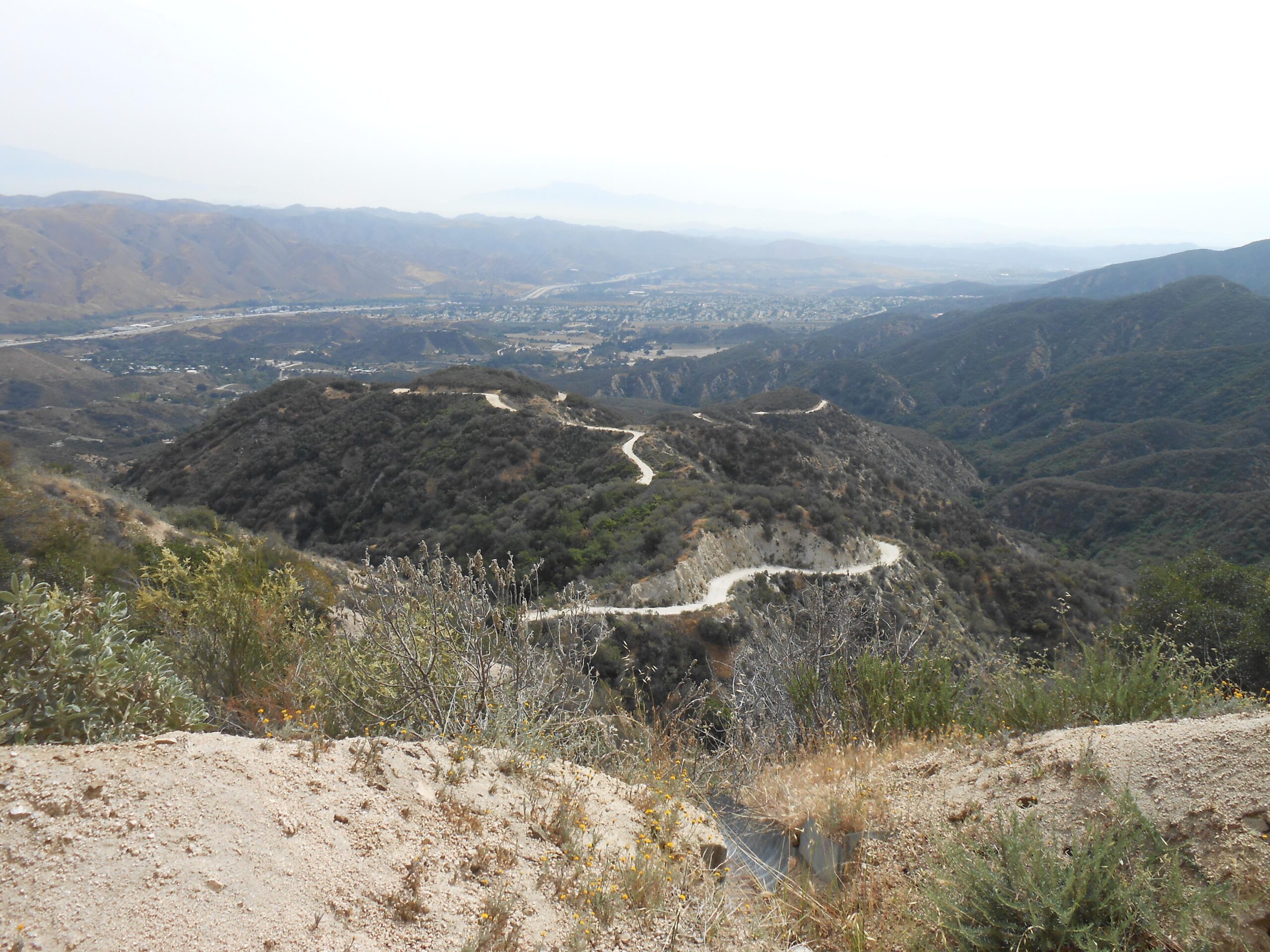 A panoramic view of rolling hills and winding dirt roads, captured from a high vantage point. In the foreground, sparse vegetation includes shrubs and grass, while the background features a vast landscape of mountains and valleys under a hazy sky. The intricate paths suggest a network of trails winding through the mountainous terrain. Indian Truck Trail mountain bike trail.