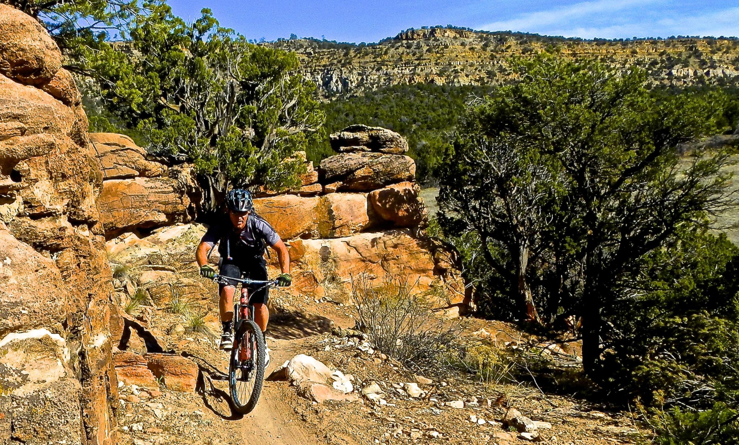 A mountain biker navigates a rocky trail through a desert landscape, surrounded by rocky outcrops and sparse vegetation, under a clear blue sky. Oil Well Flats mountain bike trail.