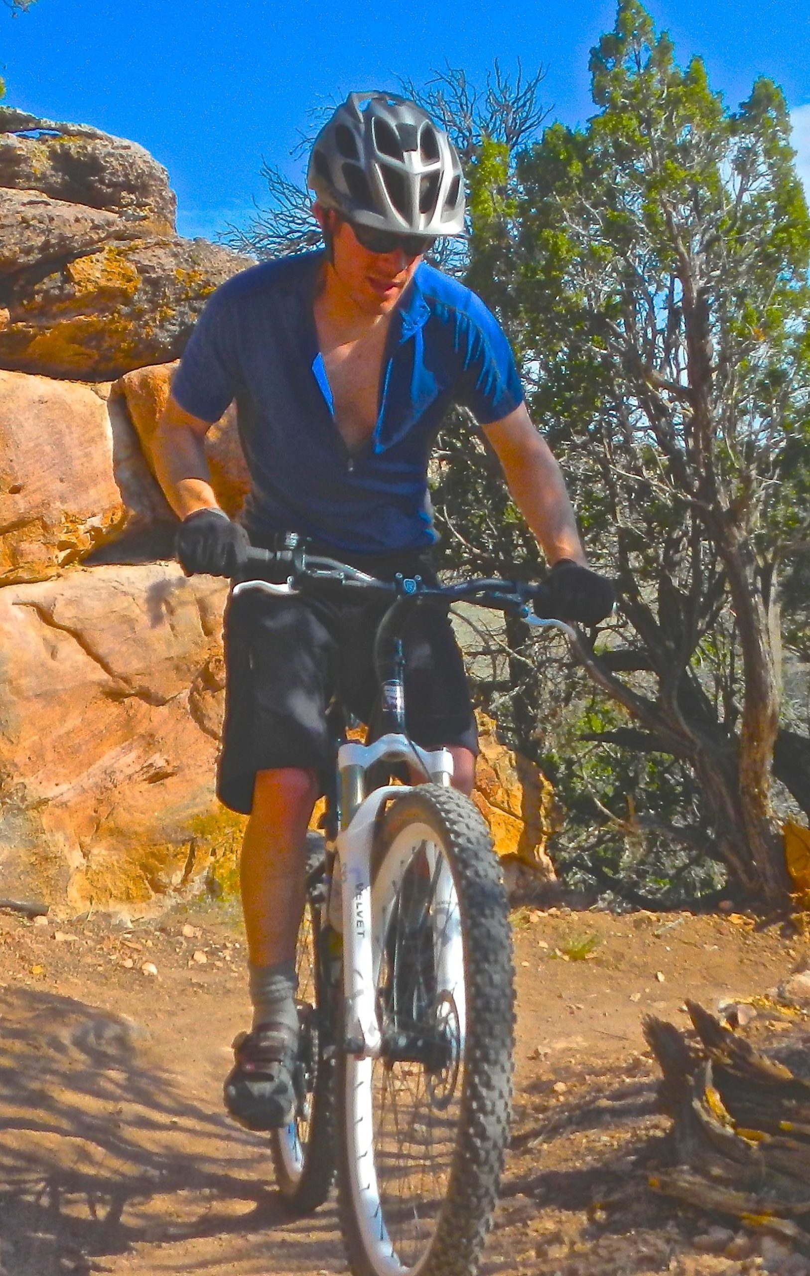 A person wearing a helmet and sunglasses rides a mountain bike on a rocky trail surrounded by trees and blue sky. They are focused on navigating the terrain, dressed in a blue shirt and black shorts. Oil Well Flats mountain bike trail.