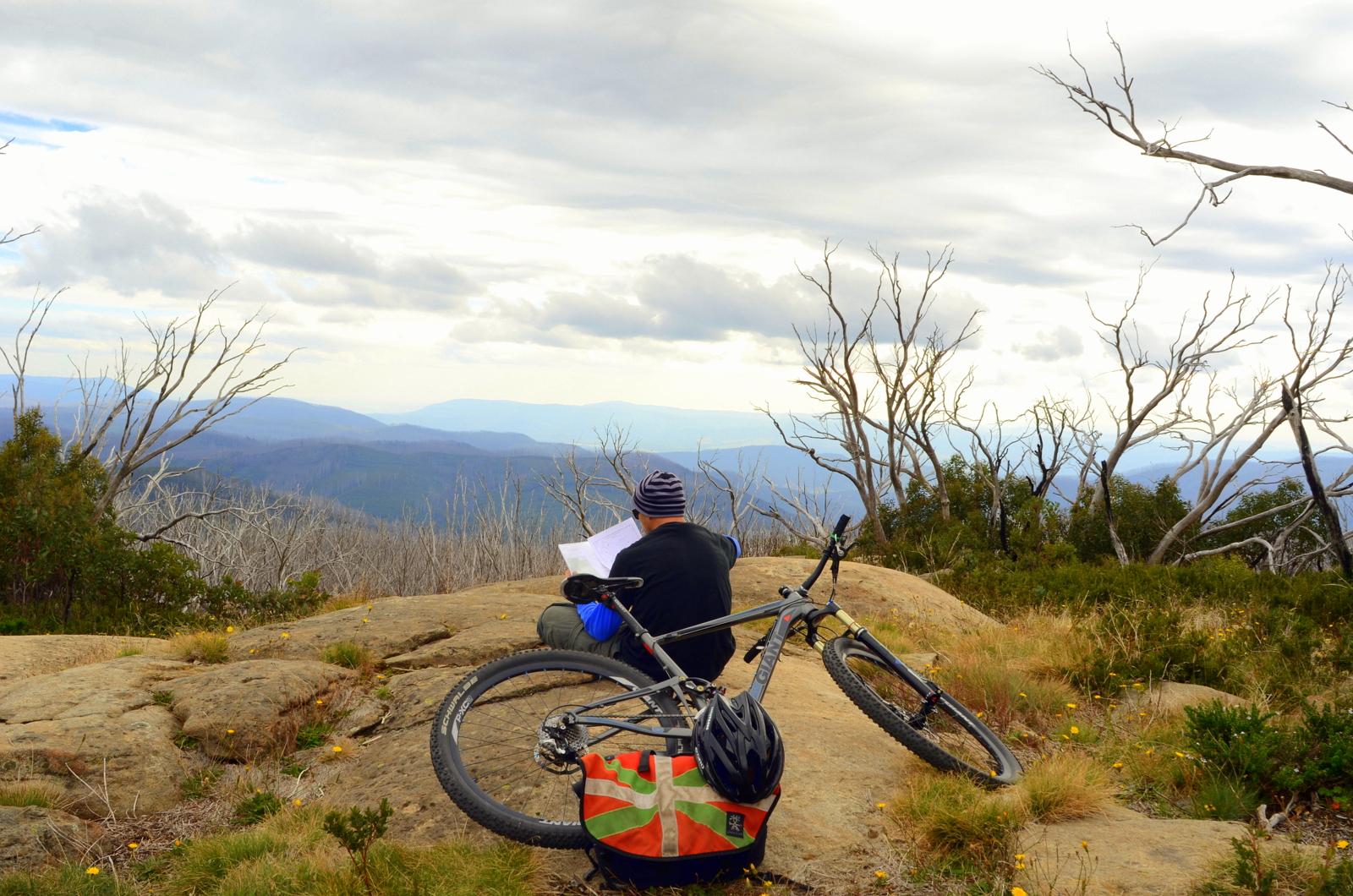 A mountain biker sits on a rock outcropping, reading a map and gazing at a scenic view of rolling mountains under a cloudy sky. A bicycle leans against the rock, and sparse trees are visible in the background, adding to the natural landscape. Lake Mountain mountain bike trail.
