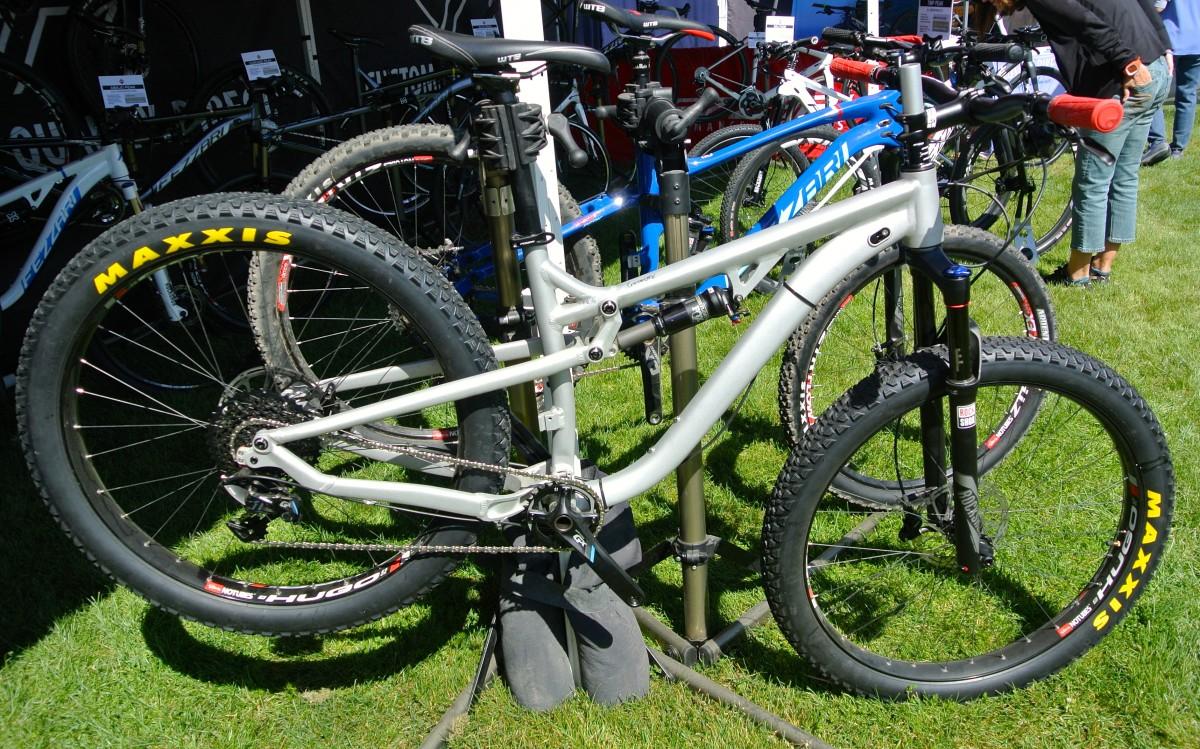 Fezzari Cascade Peak: A close-up view of a mountain bike secured on a display rack, featuring a silver frame, wide tires, and a suspension fork. In the background, several other bikes are visible, showcasing various colors and styles. The setting is outdoors on grass, indicating a bike exhibition or event.