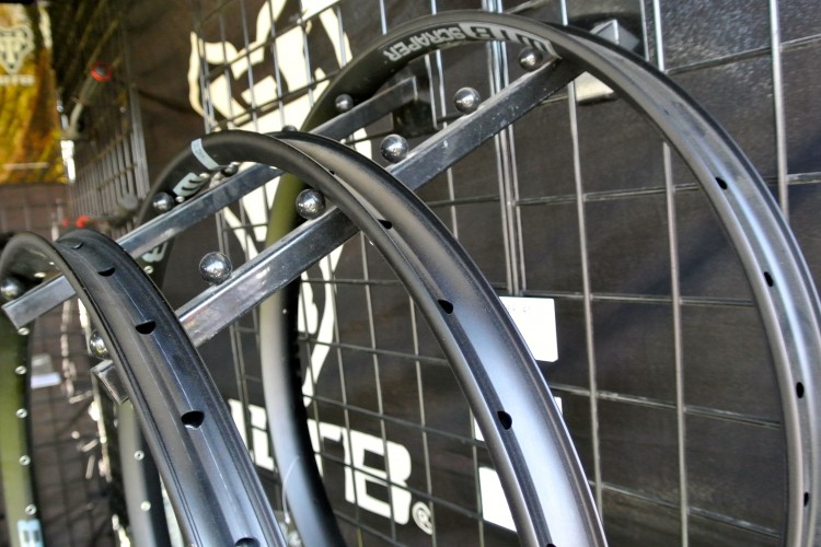 A close-up image of bike rims displayed on a rack with a grid background. The rims are circular and feature various designs and holes for spokes, showcasing different materials and finishes.