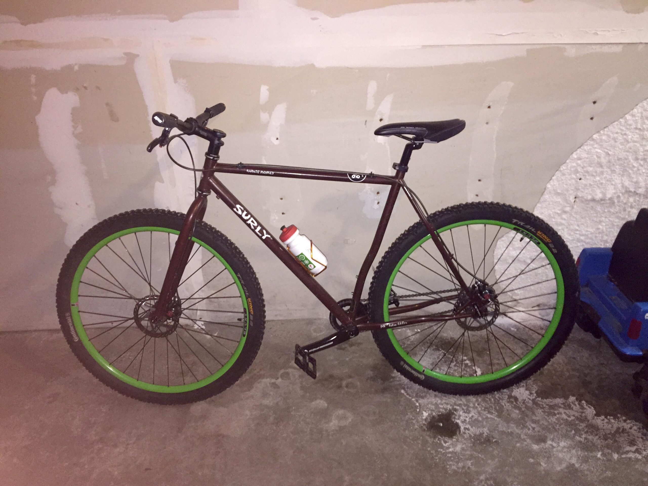 Surly Karate Monkey: A brown Surly bicycle with green rims is parked in a garage. The bike has thick tires suitable for off-road riding and a water bottle holder attached to the frame. The background features an unfinished wall with exposed patches and a blue plastic toy car.