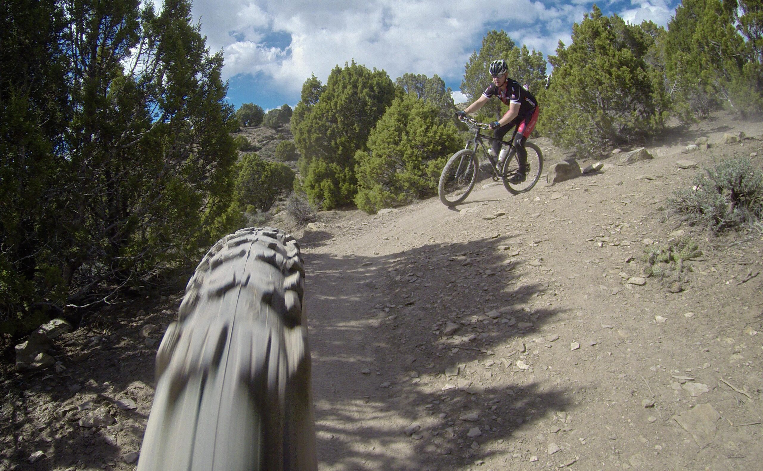 A mountain biker rides along a dirt trail surrounded by greenery, with a close-up view of the bike's front tire in motion. Dust is kicked up from the ground, and the sky is partly cloudy. Berry Creek Loop mountain bike trail.