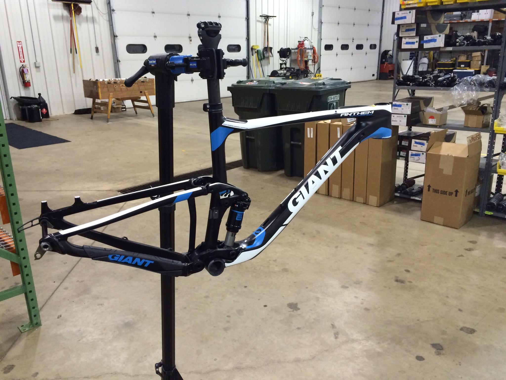 Giant Anthem Advanced 27.5 0 Team: A black and blue bike frame from the brand Giant is displayed on a repair stand in a well-lit workshop. The background features storage racks filled with bike parts and boxes, as well as various tools and equipment.