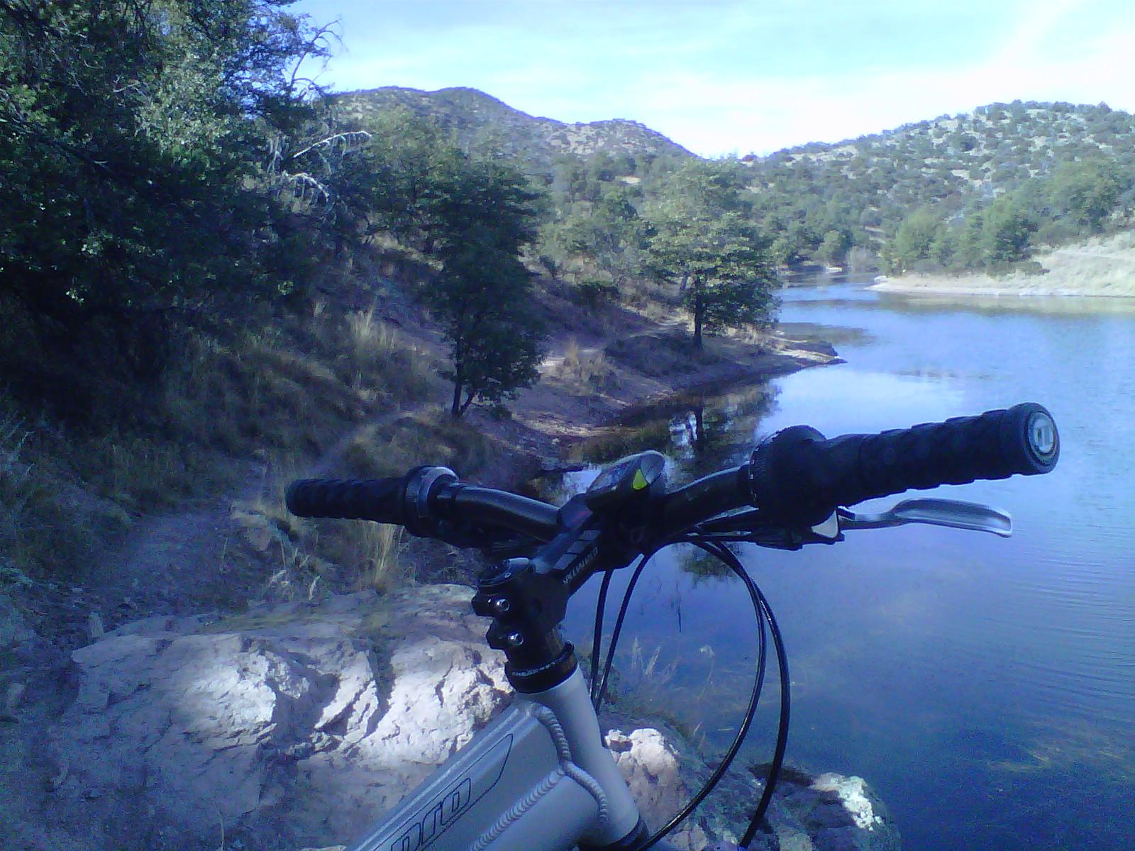 A close-up view of a mountain bike handlebar resting on a rocky edge, overlooking a serene lake surrounded by hills and trees under a clear blue sky. The scene captures the tranquility of nature, with a pathway visible leading into the greenery on the left. Parker Canyon Lake Perimeter mountain bike trail.