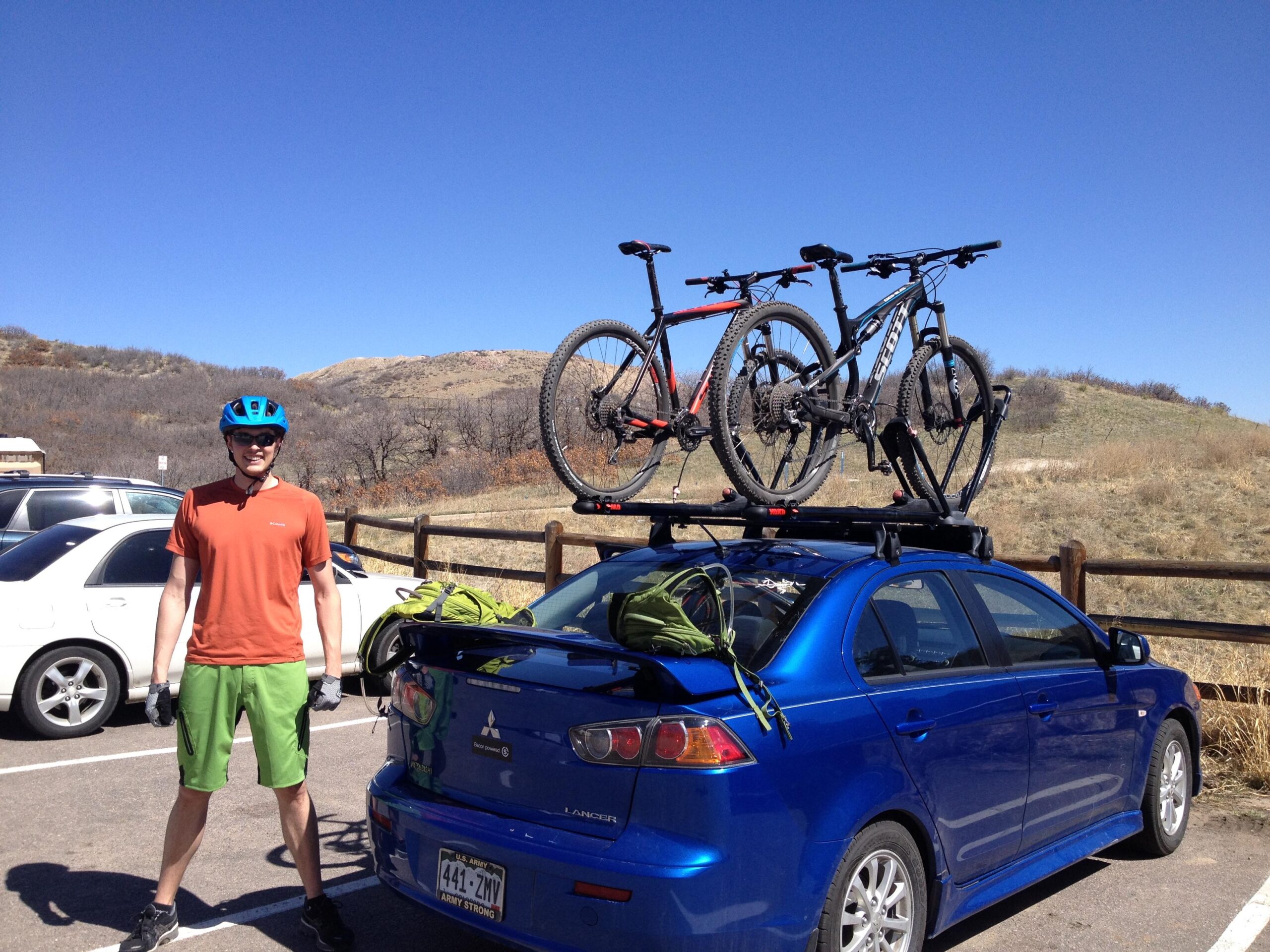 Motobecane NightTrain Bullet: A man in an orange shirt and green shorts stands next to a blue car parked in a lot. Two mountain bikes are secured on the roof rack of the car. The background features a clear blue sky and a hillside with sparse vegetation.