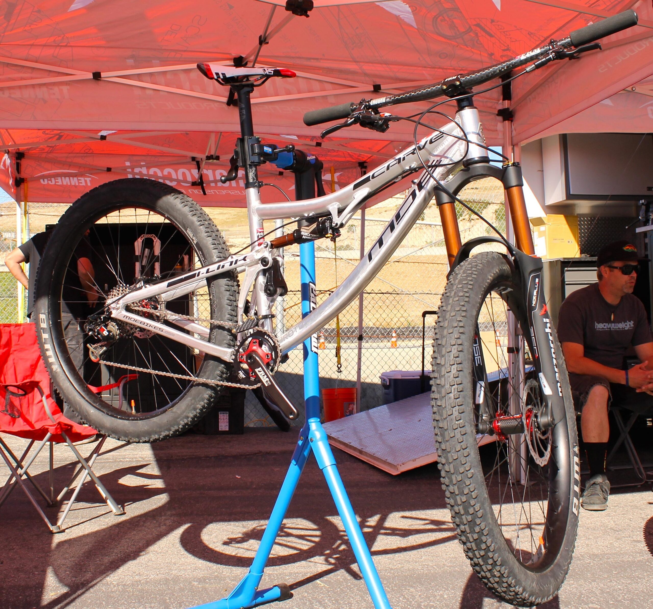 MDE Carve: A silver mountain bike suspended on a blue repair stand, with prominent features like wide tires and a front fork. In the background, a tent with branding is set up, and a person is seated nearby, partially visible. The scene is set in an outdoor environment with a slight incline.