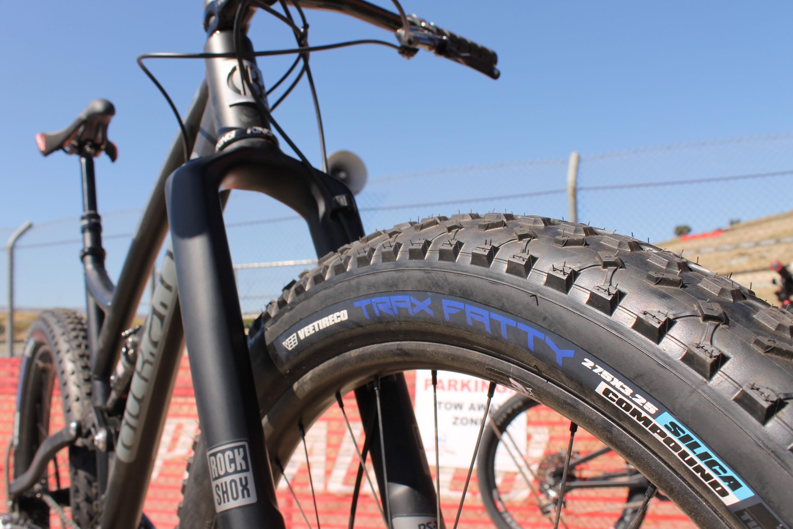 Durango Bike Company Blackjack: Close-up view of a mountain bike's front tire and fork, featuring a wide tread pattern for off-road traction. The tire is labeled "Trax Fatty," and the fork bears the "Rock Shox" logo. The background includes a fence and a parking sign, with a clear blue sky above.