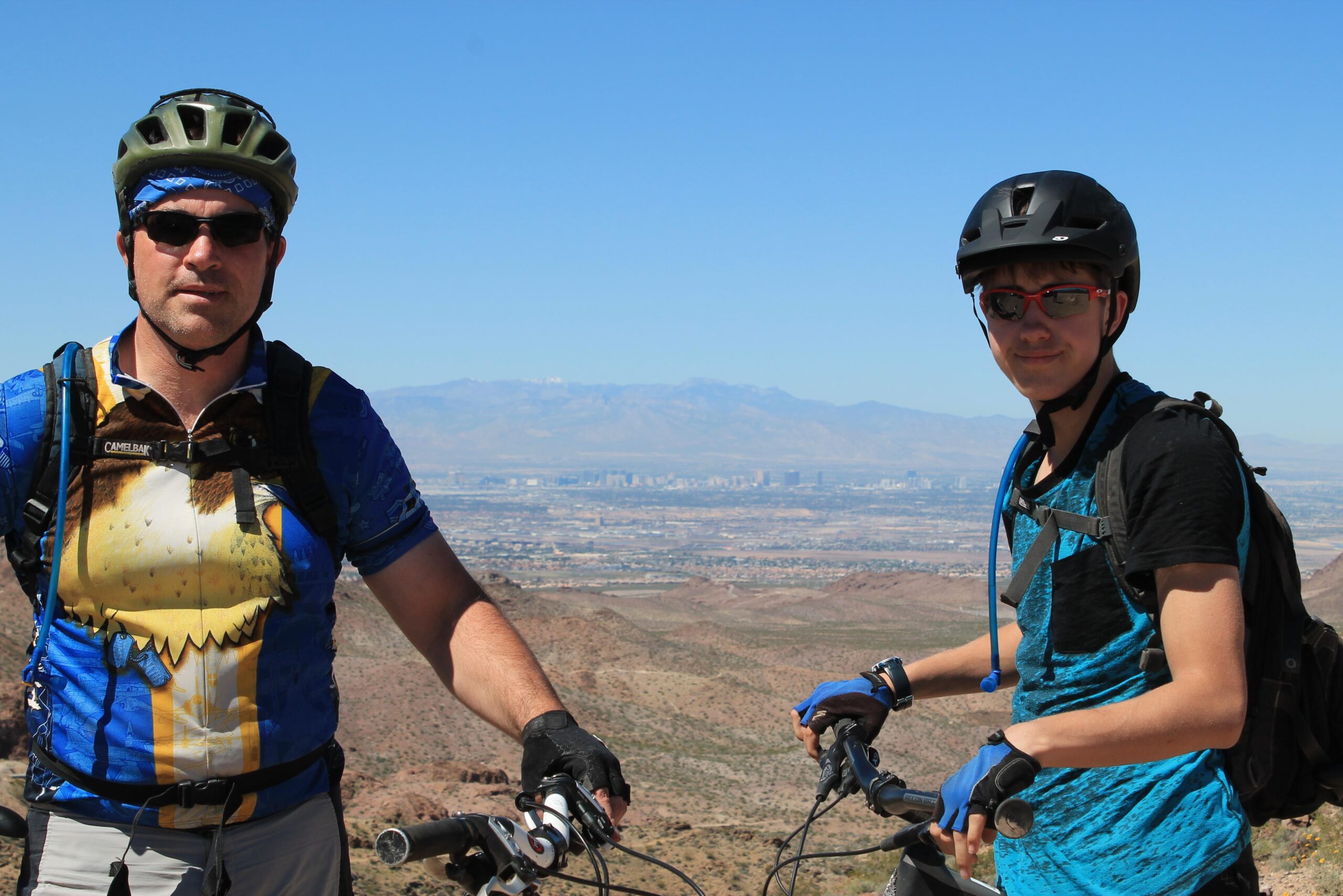 Two mountain bikers pose for a photo on a scenic overlook, showcasing a panoramic view of a valley. The adult is wearing a colorful cycling jersey and a helmet, while the younger cyclist is in a blue shirt and a black helmet. Both are holding their bike handlebars. The background features mountains and a clear blue sky. Bootleg Canyon mountain bike trail.