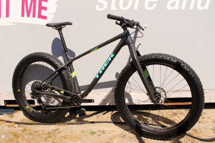 A black Trek mountain bike with large tires, featuring a sleek design and vibrant blue and yellow accents, positioned against a light gray backdrop with text that reads "Try Me" and "store and." The bike is displayed in profile, emphasizing its frame and wheel size.