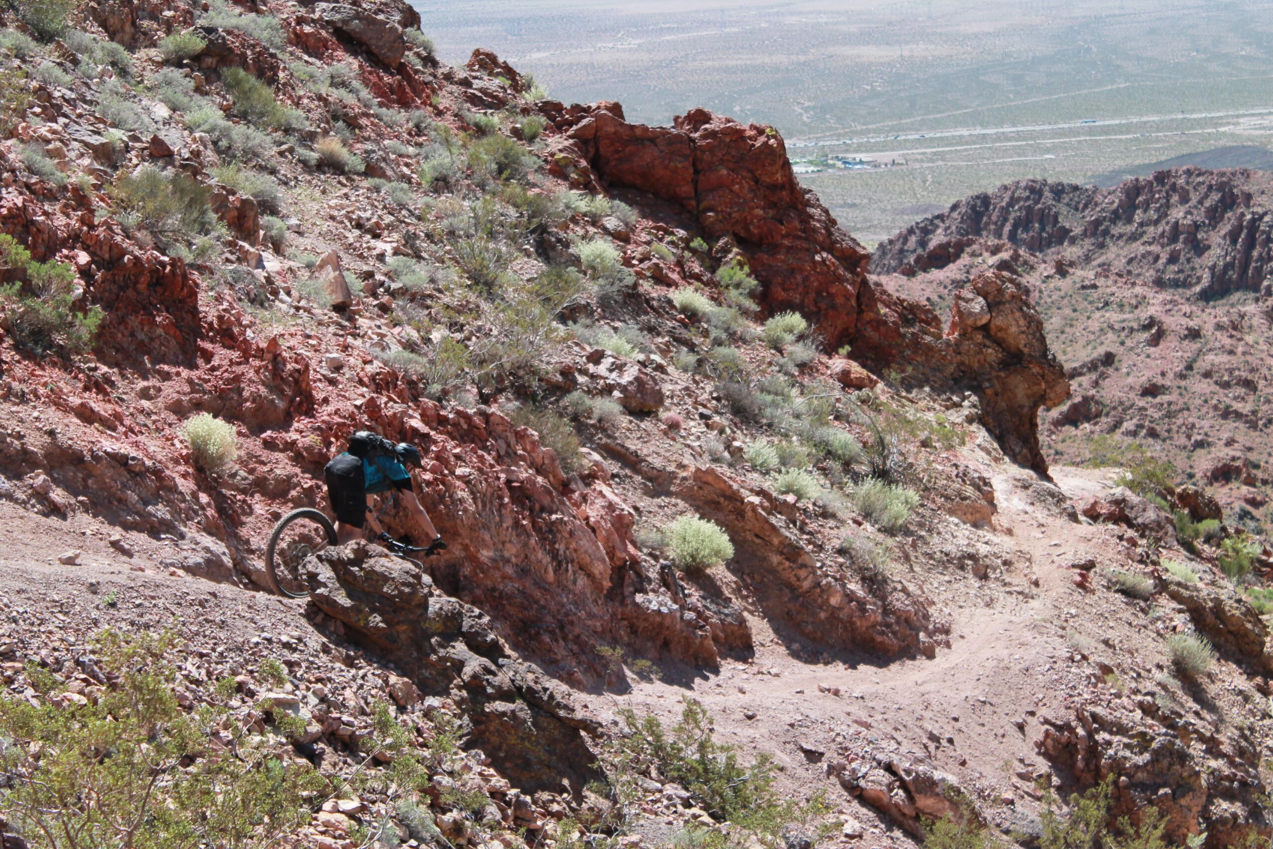 A cyclist navigates a rocky mountain trail surrounded by red rock formations and sparse vegetation in a desert landscape. The winding path leads into the distance, showcasing a rugged, scenic terrain. Bootleg Canyon mountain bike trail.