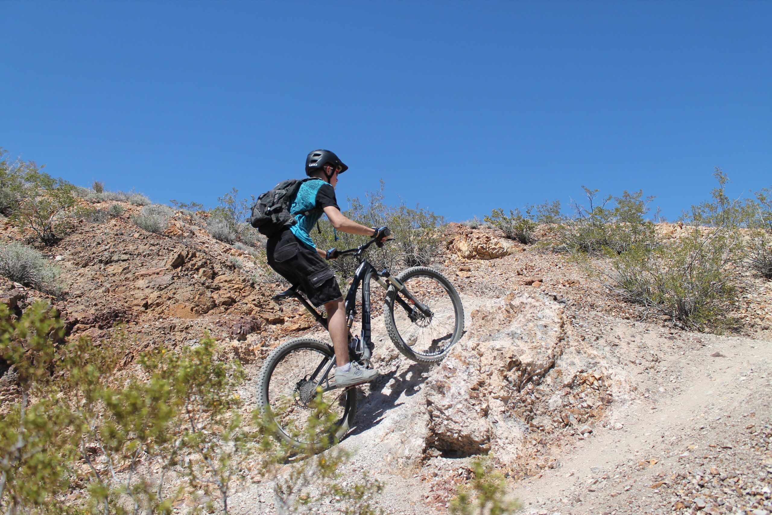 A person mountain biking uphill on rocky terrain in a sunny, arid landscape. The rider is wearing a helmet and a blue and black shirt, with a backpack, navigating through sparse vegetation and rocky ground under a clear blue sky. Bootleg Canyon mountain bike trail.
