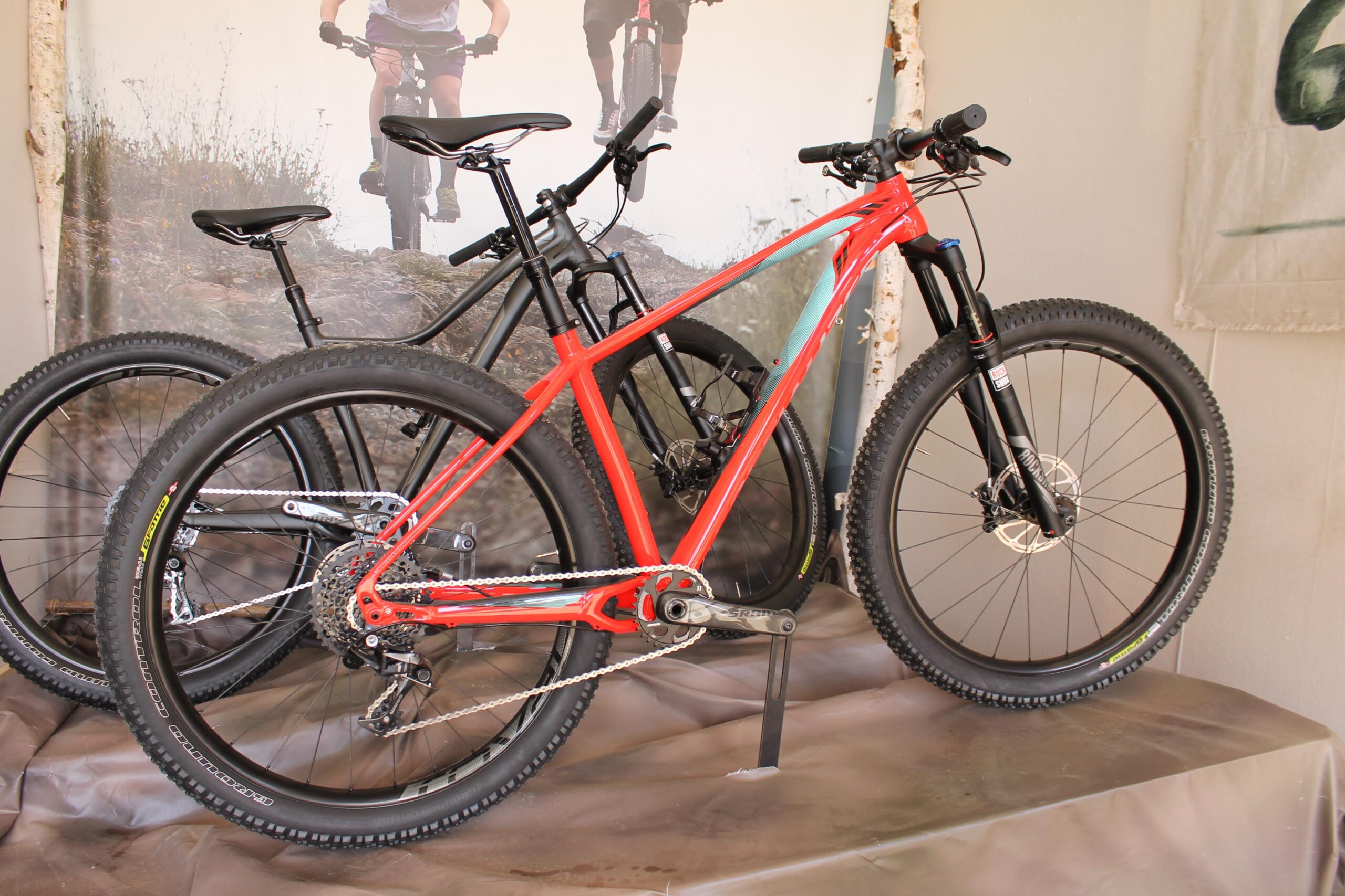 Specialized Fuse: A vibrant red mountain bike is prominently displayed in the foreground, showcasing its sleek frame and thick tires. Behind it are two additional bikes, one in black and another partially visible, all set against a backdrop featuring outdoor cycling scenery. The display is arranged on a neutral-colored surface.