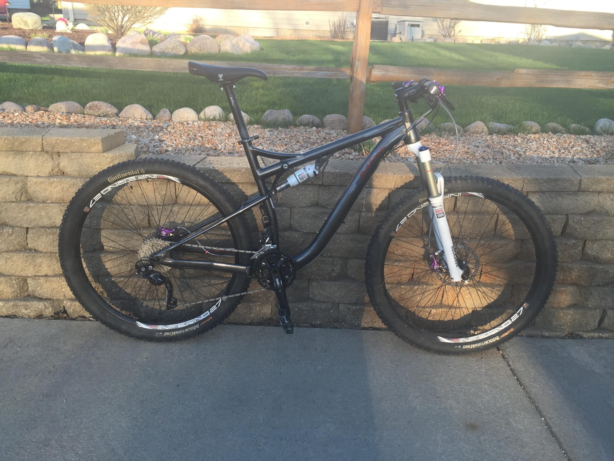 Salsa Horsethief: Alt text: A black mountain bike with large tires and a white front fork, positioned against a stone retaining wall. The background features a grassy area and a wooden fence.