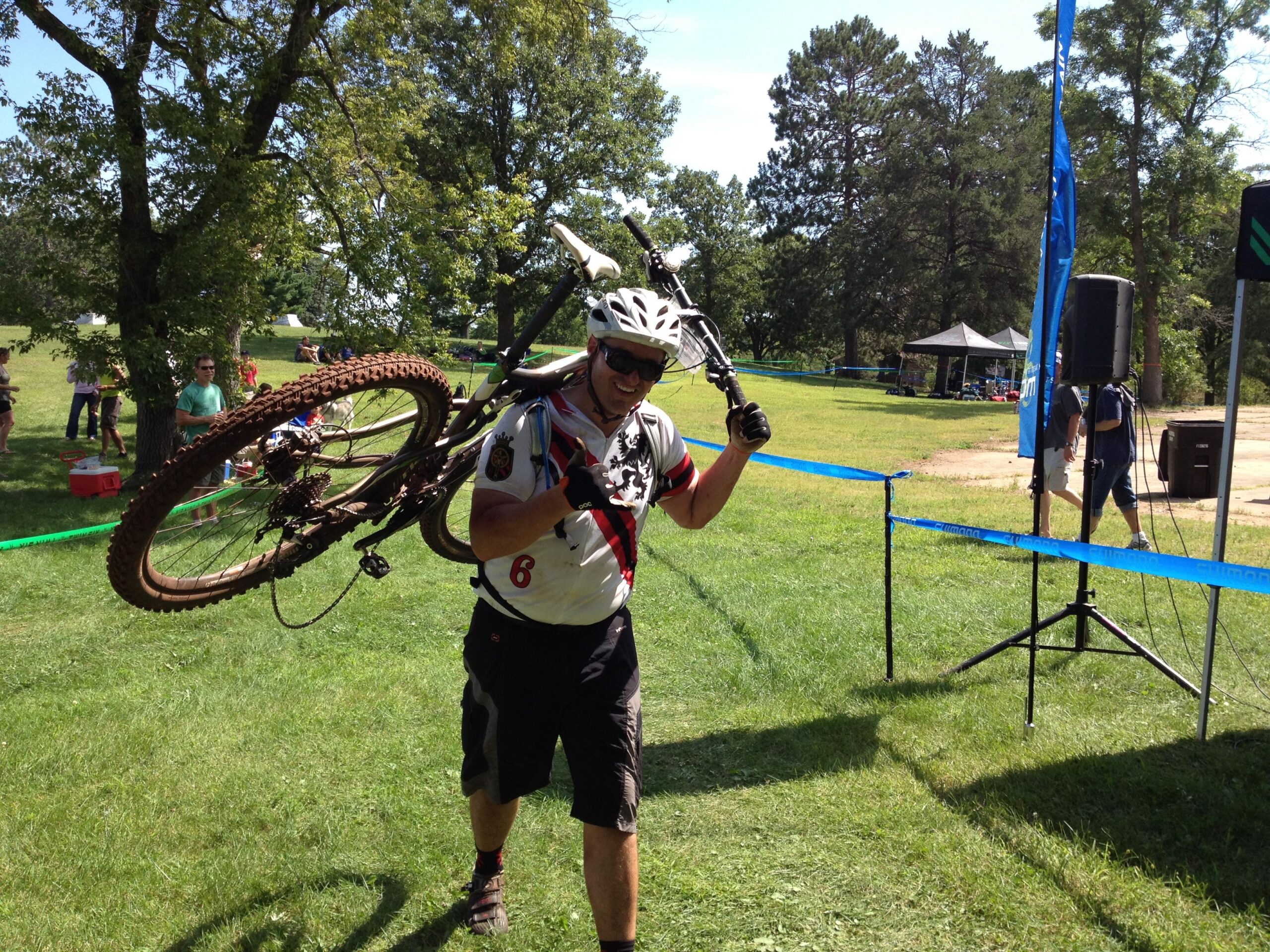 Motobecane NightTrain Bullet: A cyclist wearing a helmet and a jersey carrying a mountain bike on their shoulder while walking through a grassy area. In the background, event tents and spectators can be seen enjoying the outdoor setting on a sunny day.