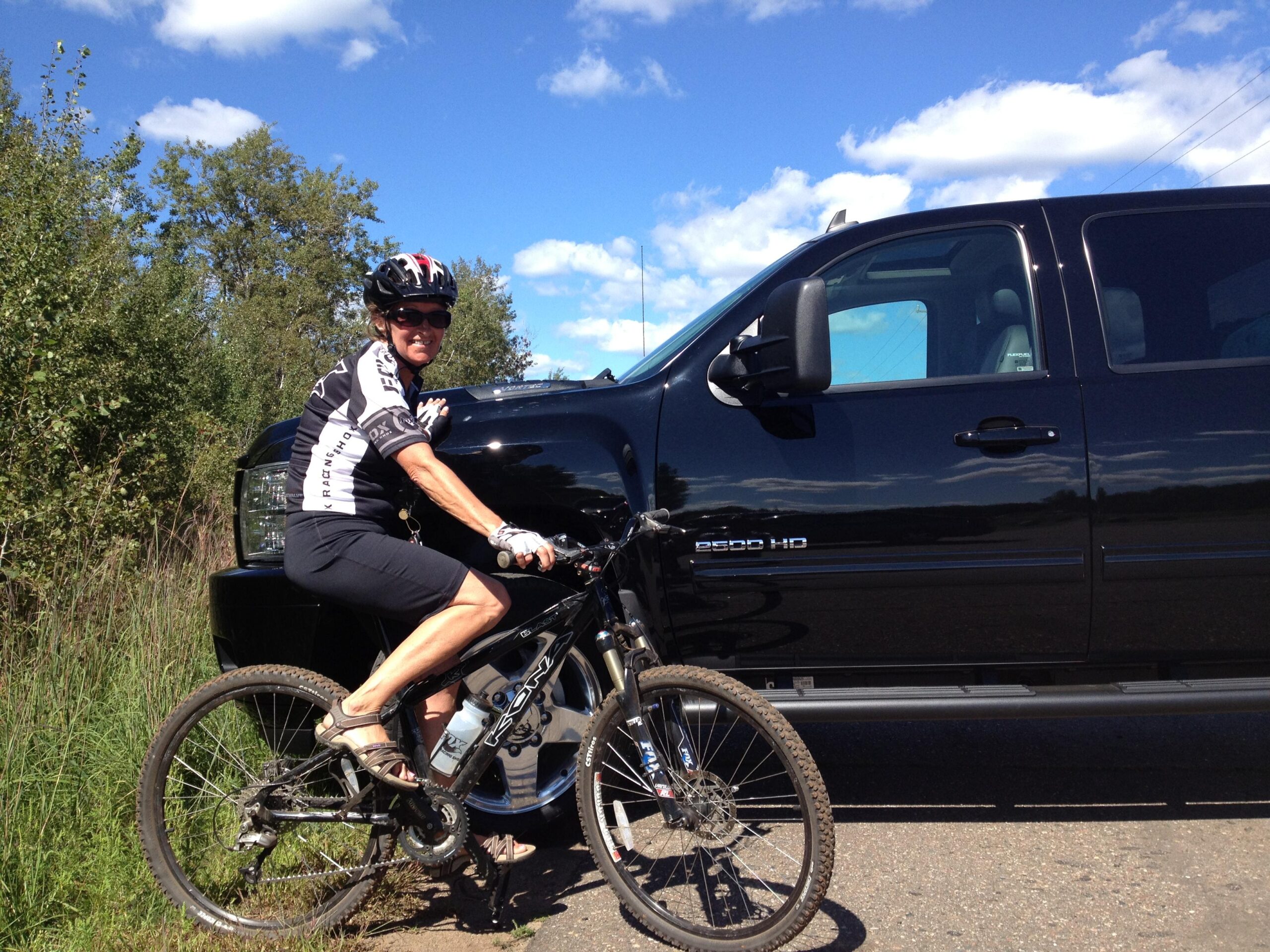 Motobecane NightTrain Bullet: A cyclist wearing a black and white jersey and a helmet is leaning against a black pickup truck, with a backdrop of blue skies and greenery. The cyclist is seated on a mountain bike, and the truck displays the "2500 HD" badge.