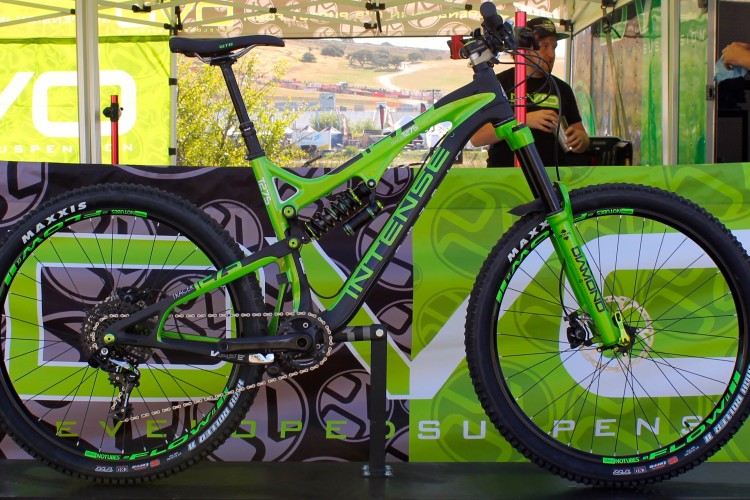 A green and black mountain bike displayed in front of a branded backdrop at an outdoor event. The bike features large tires, a dual suspension system, and various components labeled with brand names. The scene includes a few people in the background engaging with the event setup.