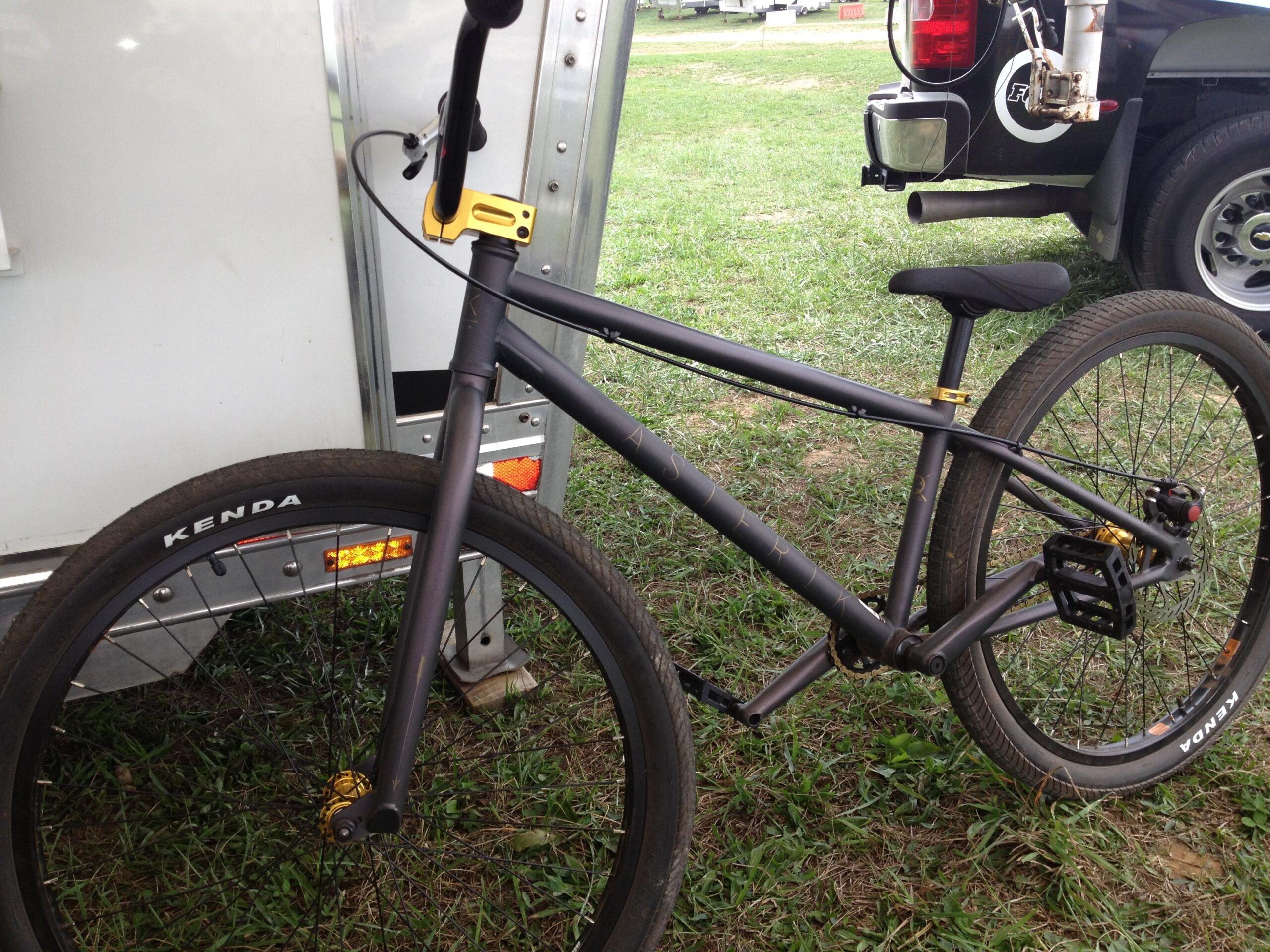 Motobecane NightTrain Bullet: A black BMX bike with yellow accents parked on grass next to a silver trailer. The bike features a sturdy frame, thick tires with "Kenda" branding, and a simple seat, highlighting its design for performance and tricks.