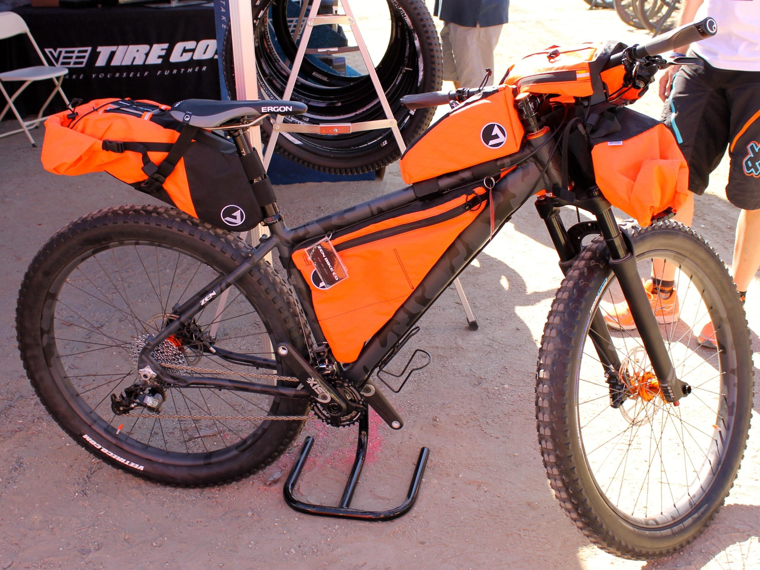 Zen 27.5: A mountain bike presented with orange and black bags for storage, showcasing a rugged design suitable for outdoor adventures. The bike is displayed on a gravel surface, with a background hinting at a bike event or expo setting.