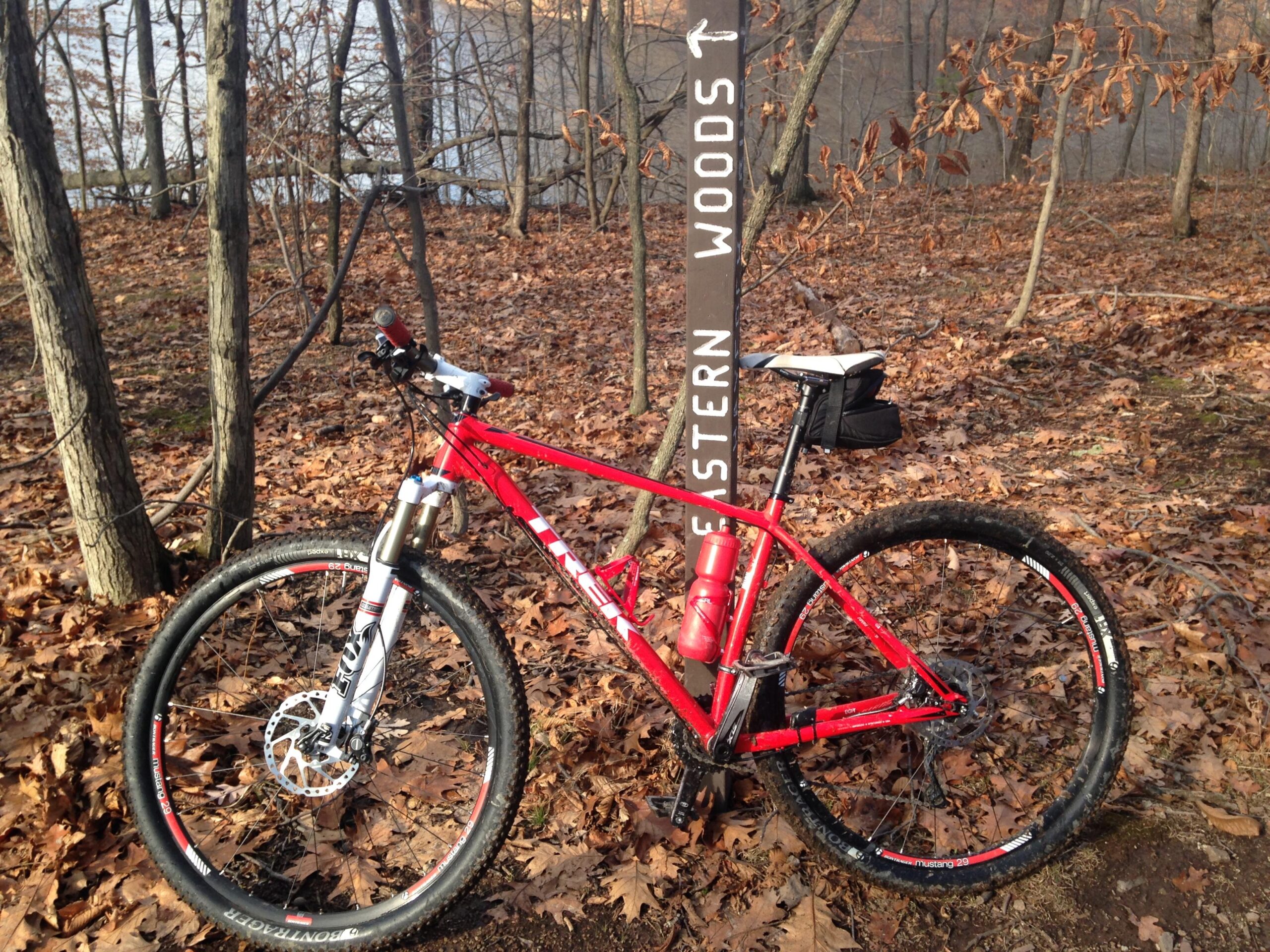 Motobecane NightTrain Bullet: A red mountain bike is parked beside a trail sign that reads "Eastern Woods." The bike features front suspension and has a water bottle attached to the frame. The background shows a wooded area with bare trees and scattered brown leaves.