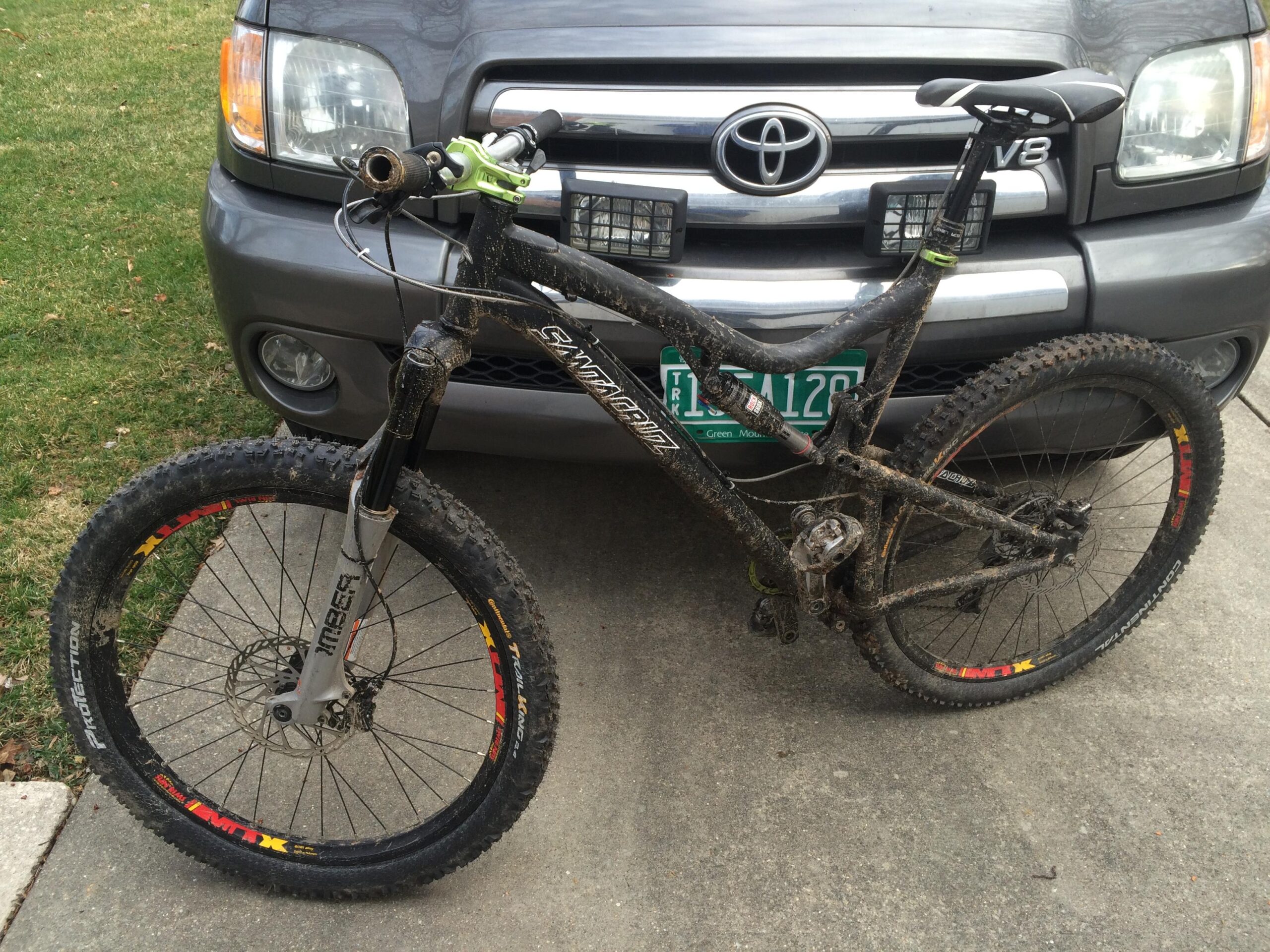 Santa Cruz Blur LT2: A mountain bike with a dirty frame and wheels, parked on a concrete driveway in front of a gray Toyota SUV. The bike features thick tires and a dual-suspension setup. The environment includes a patch of grass in the background.
