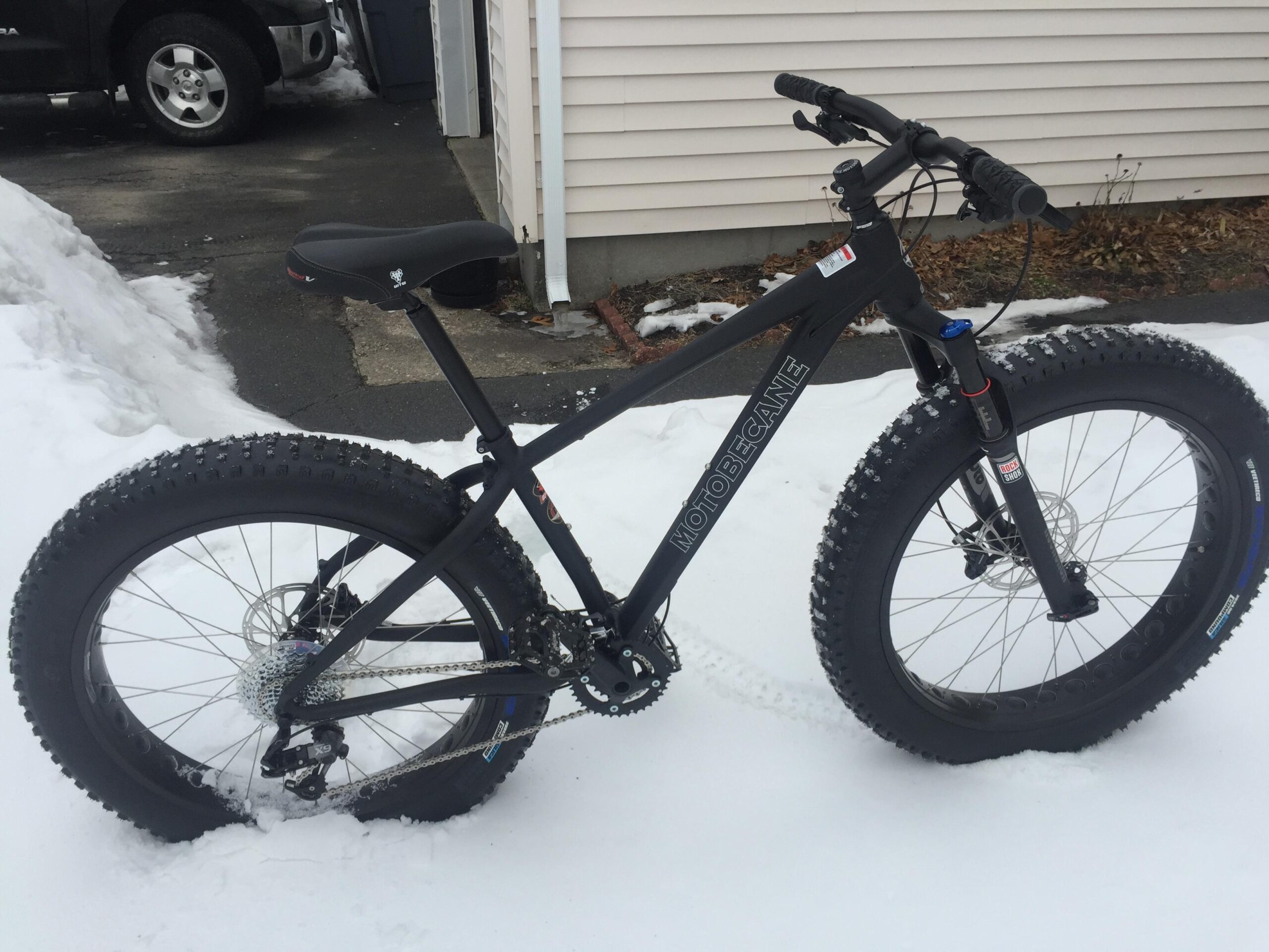 Motobecane NightTrain Bullet: A fat tire bike parked on snow-covered ground, featuring a black frame and thick, knobby tires designed for winter riding. In the background, a glimpse of a driveway and a house is visible, suggesting a suburban setting.