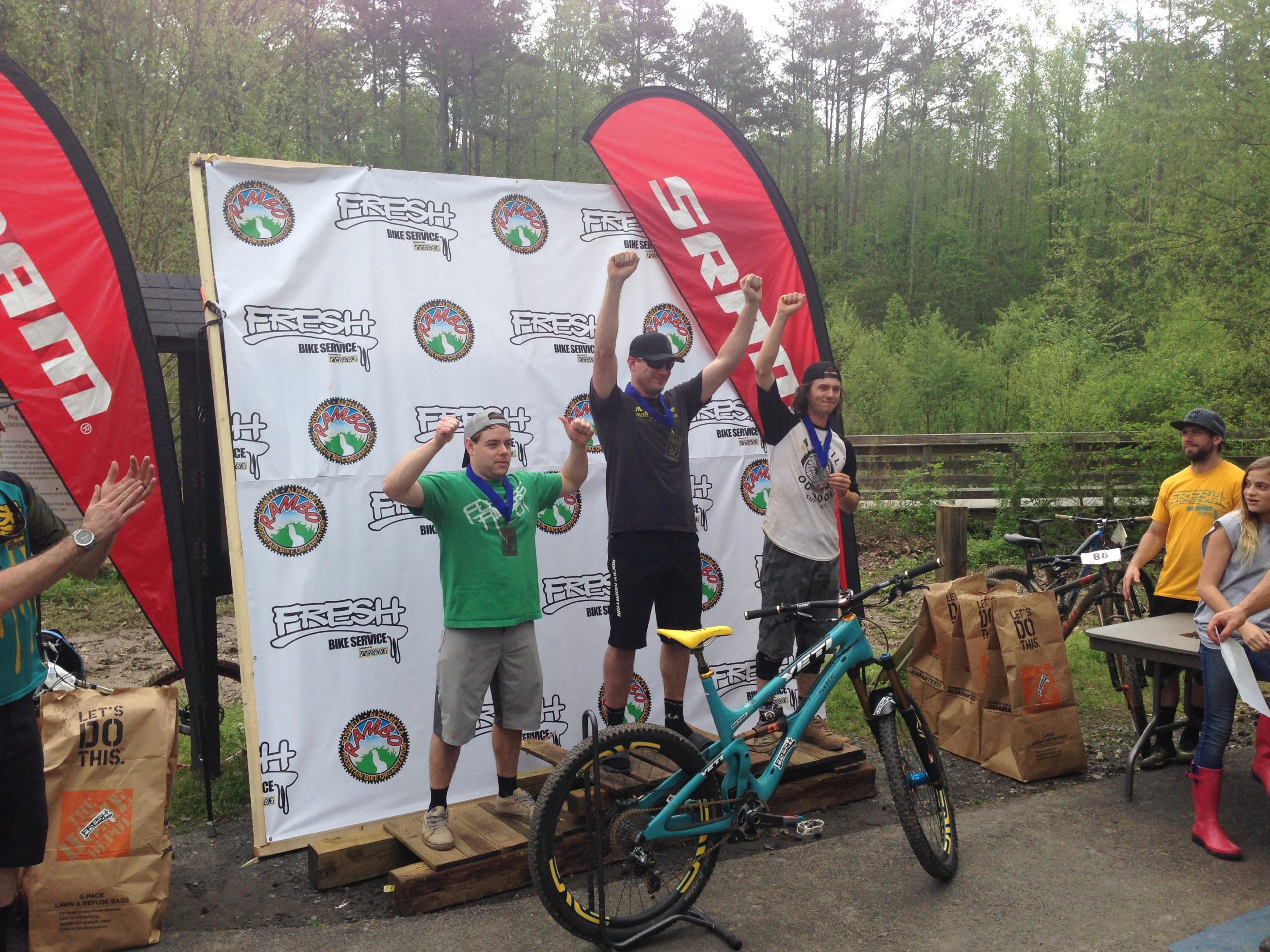 Three male mountain bikers celebrate on a podium, raising their arms in victory while holding medals. The two individuals on the podium wear casual athletic clothing, with one in a green shirt and the other in a white shirt. In the background, a backdrop displays logos for "Fresh Bike Service" and a forested area is visible. Two additional spectators observe and applaud from the side, with a table displaying papers nearby. An off-road bicycle is positioned on the podium, and bags labeled "Let's Do This" are seen on the ground. Big Creek mountain bike trail.