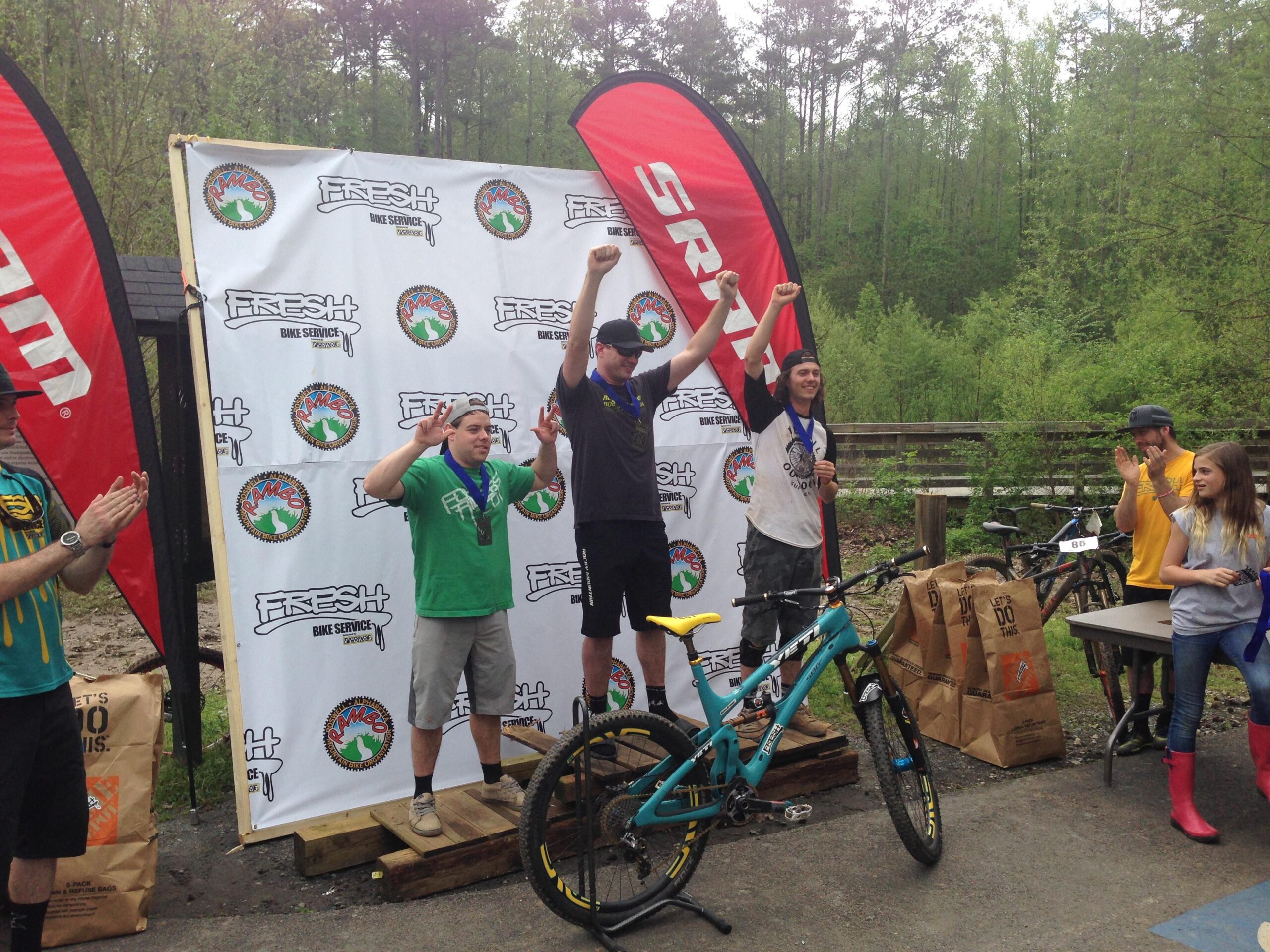 Three male mountain bikers celebrate on a podium after a race, with one holding a medal and two raising their fists in victory. They are surrounded by a cheering crowd, including a young girl and other participants. The backdrop features banners from the event sponsor, and mountain bikes are placed nearby. The setting is a wooded area, indicating an outdoor biking competition. Big Creek mountain bike trail.