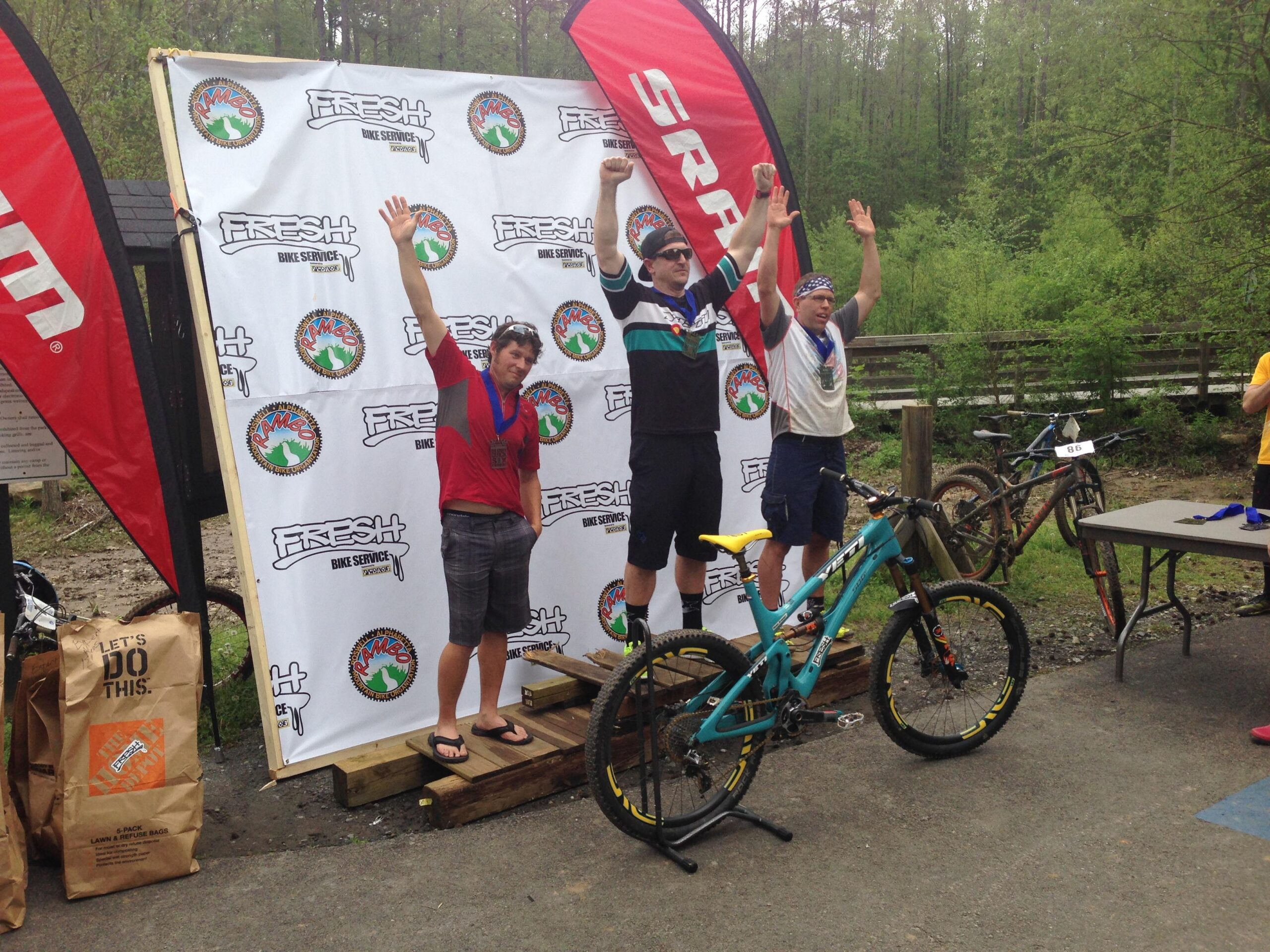Three male cyclists stand on a podium celebrating their achievements after a race. The first place winner is in the center, wearing a medal and raising his hands in victory. The second place finisher is on the left, also wearing a medal and smiling. The third place finisher is on the right, with arms raised and a joyful expression. In the background, there is a branded backdrop featuring logos, and bicycles are parked nearby. Big Creek mountain bike trail.