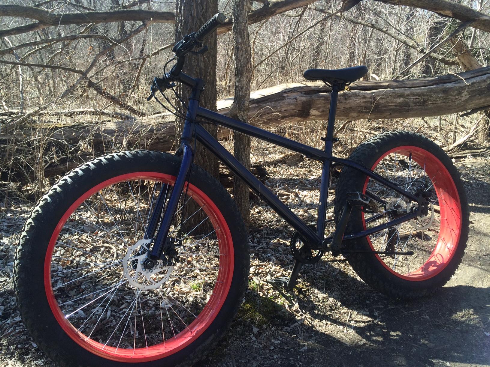 Mongoose Dolomite: A blue mountain bike with large, red tires parked beside a tree in a wooded area. The ground is covered with leaves and twigs, and a fallen log is visible in the background, indicating a natural trail setting.