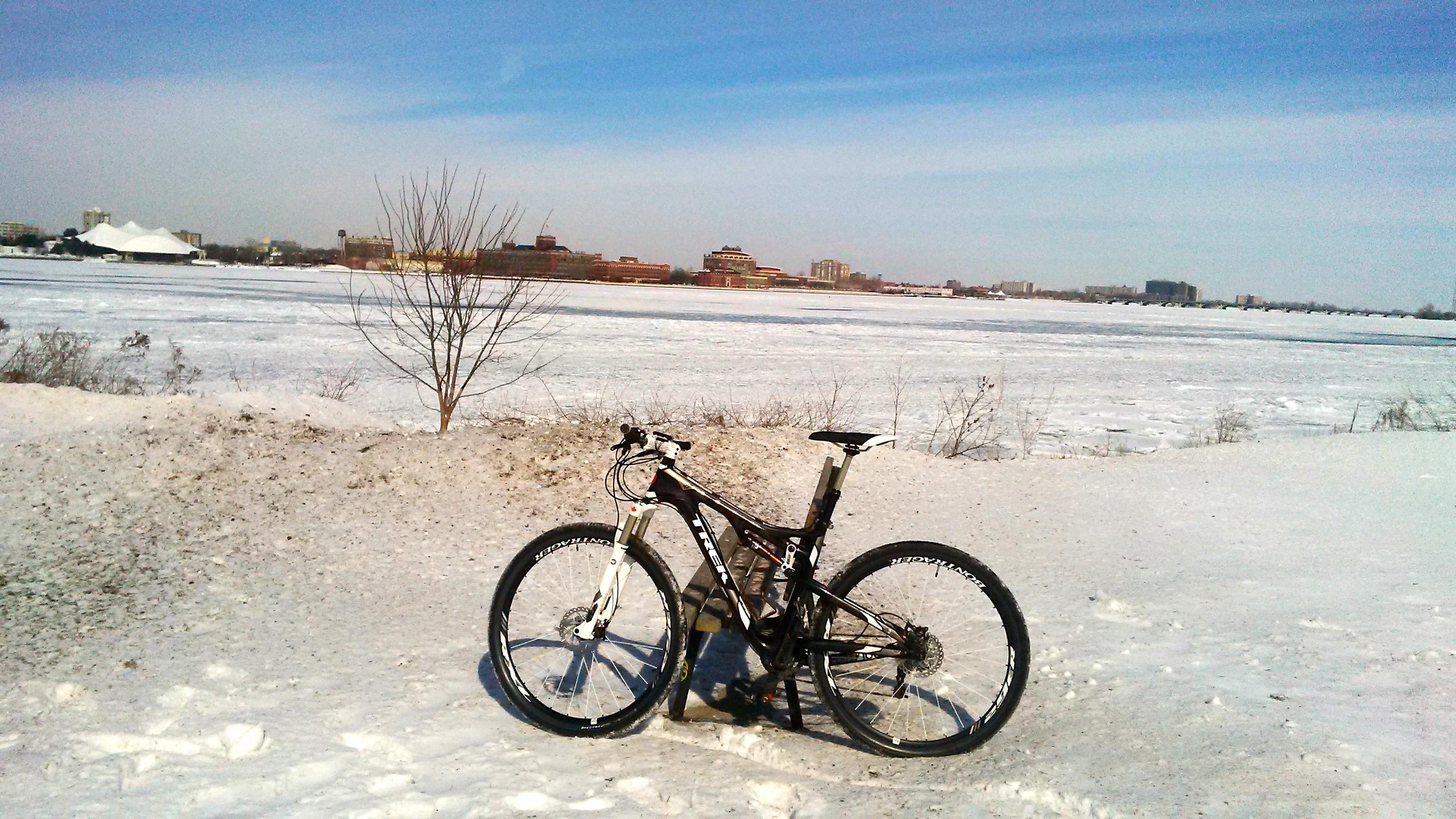Motobecane NightTrain Bullet: A black mountain bike is parked on a snowy landscape beside a frozen body of water, with urban buildings visible in the background under a clear blue sky.