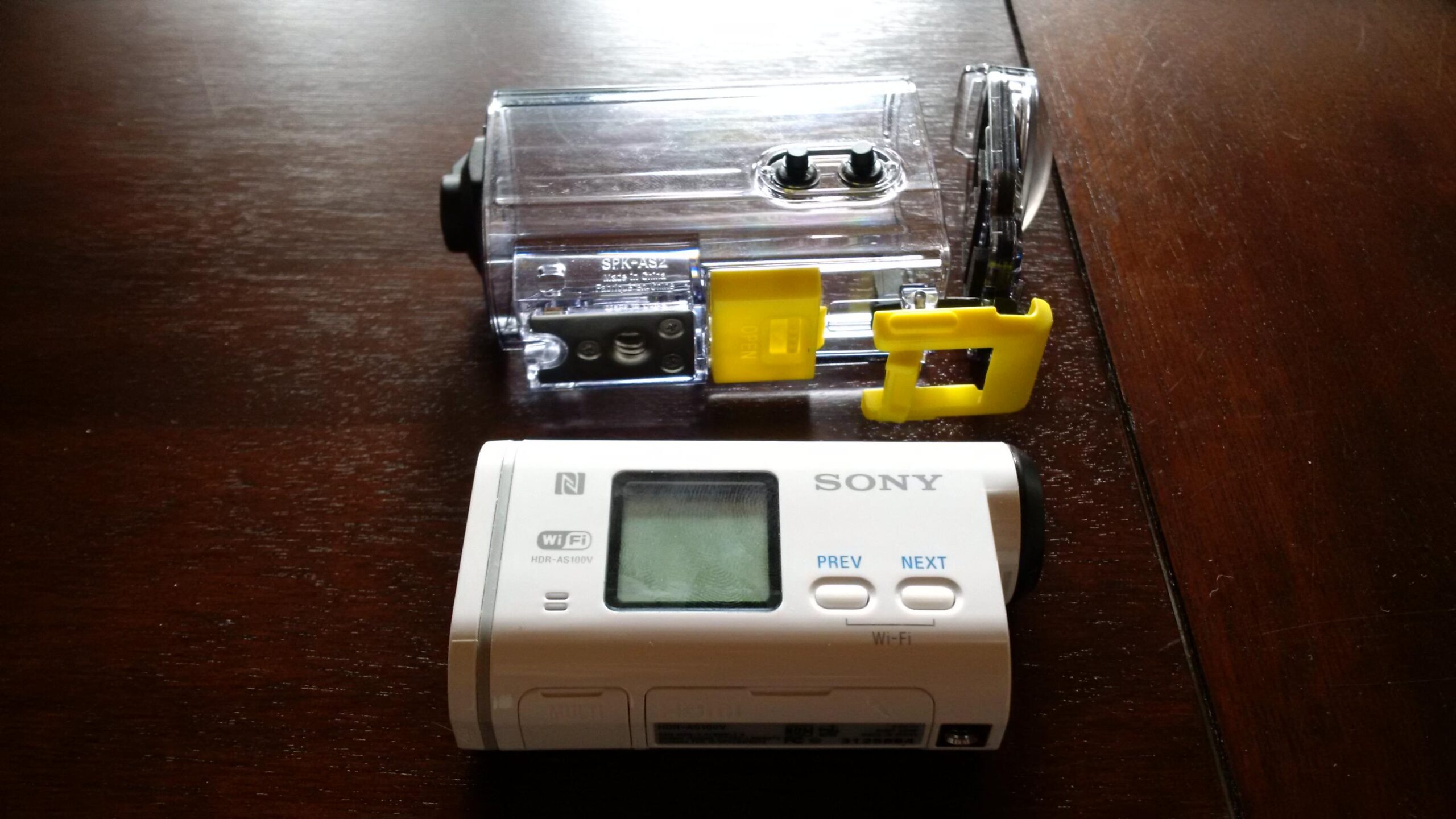 Sony HDR-AS100VR: A Sony action camera placed on a wooden surface, next to its clear waterproof housing case with a yellow latch. The camera features a small display and buttons for navigation.
